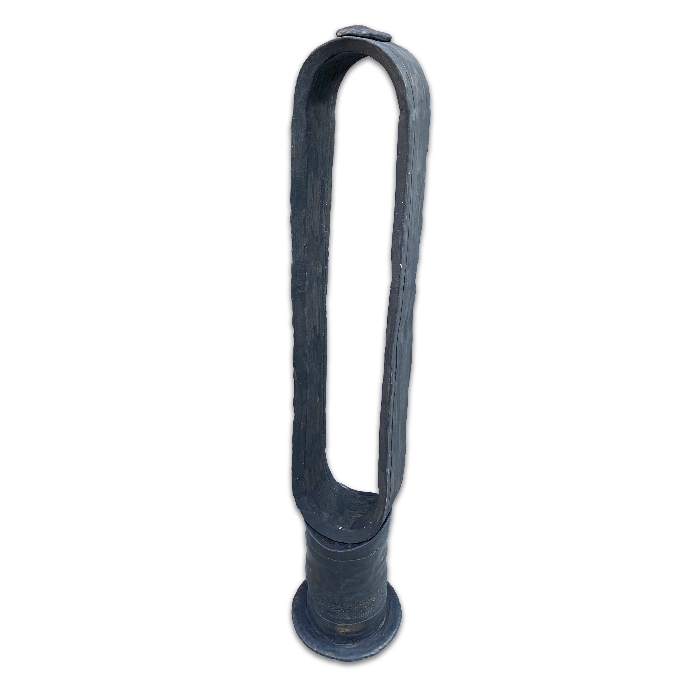 A tall, narrow sculptural form with a cylindrical base and elongated loop top, rendered in dark blue-gray glazed clay with a matte finish. The closed loop creates a minimalist, abstract silhouette against a white background.