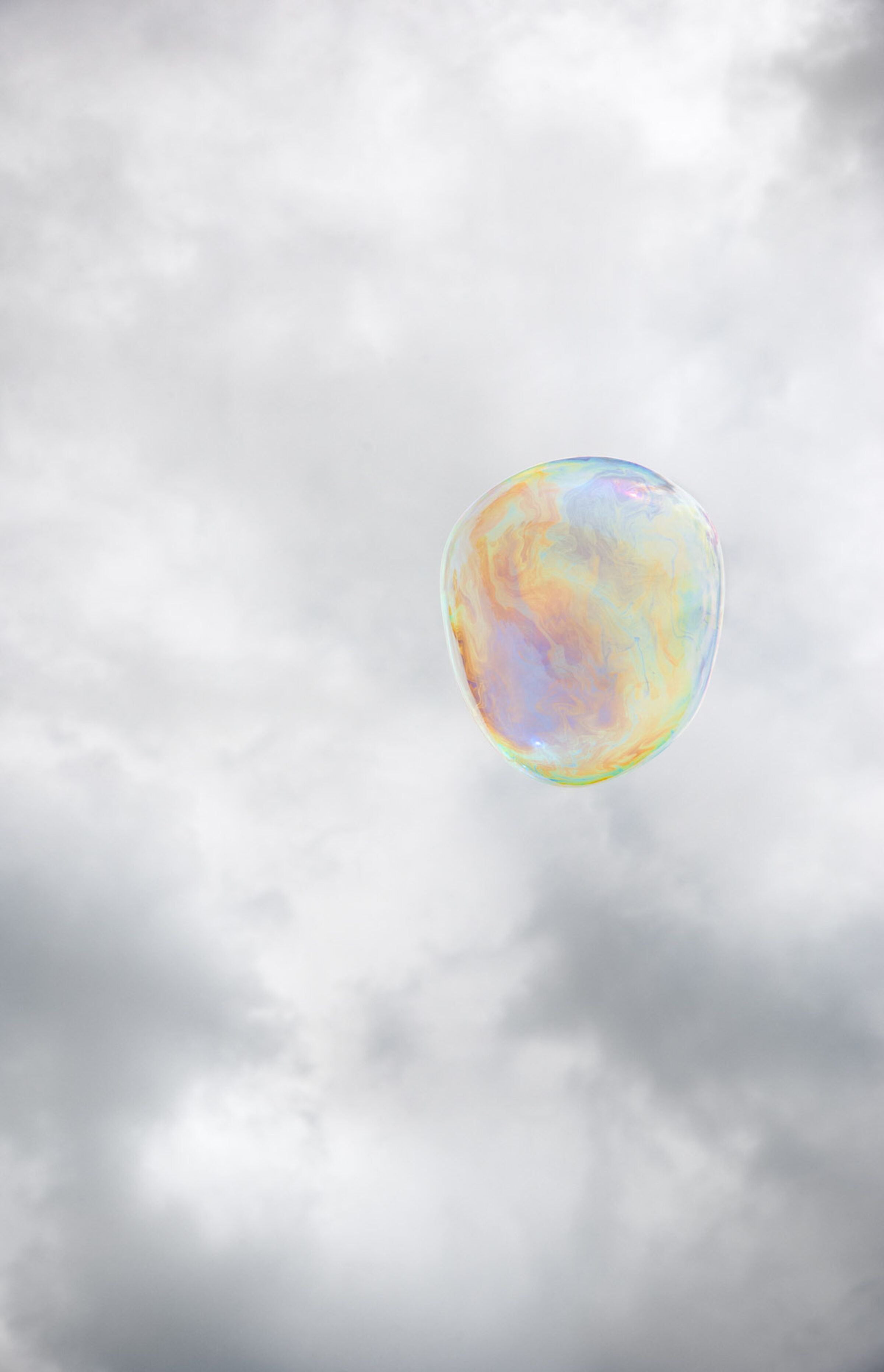 A translucent soap bubble floats against a soft, cloudy gray sky. Its spherical surface displays iridescent swirls of pastel yellow, pink, blue, and green that shift across the membrane.