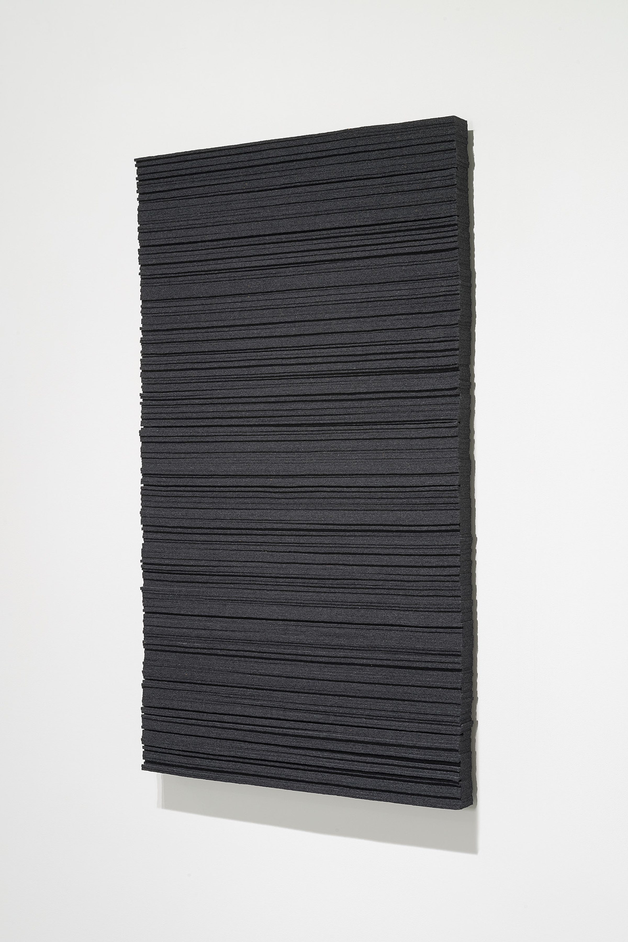 A rectangular panel of dark charcoal wool felt with horizontal linear ridges across its surface, mounted at a slight angle against a light gray background with a pale base visible beneath.