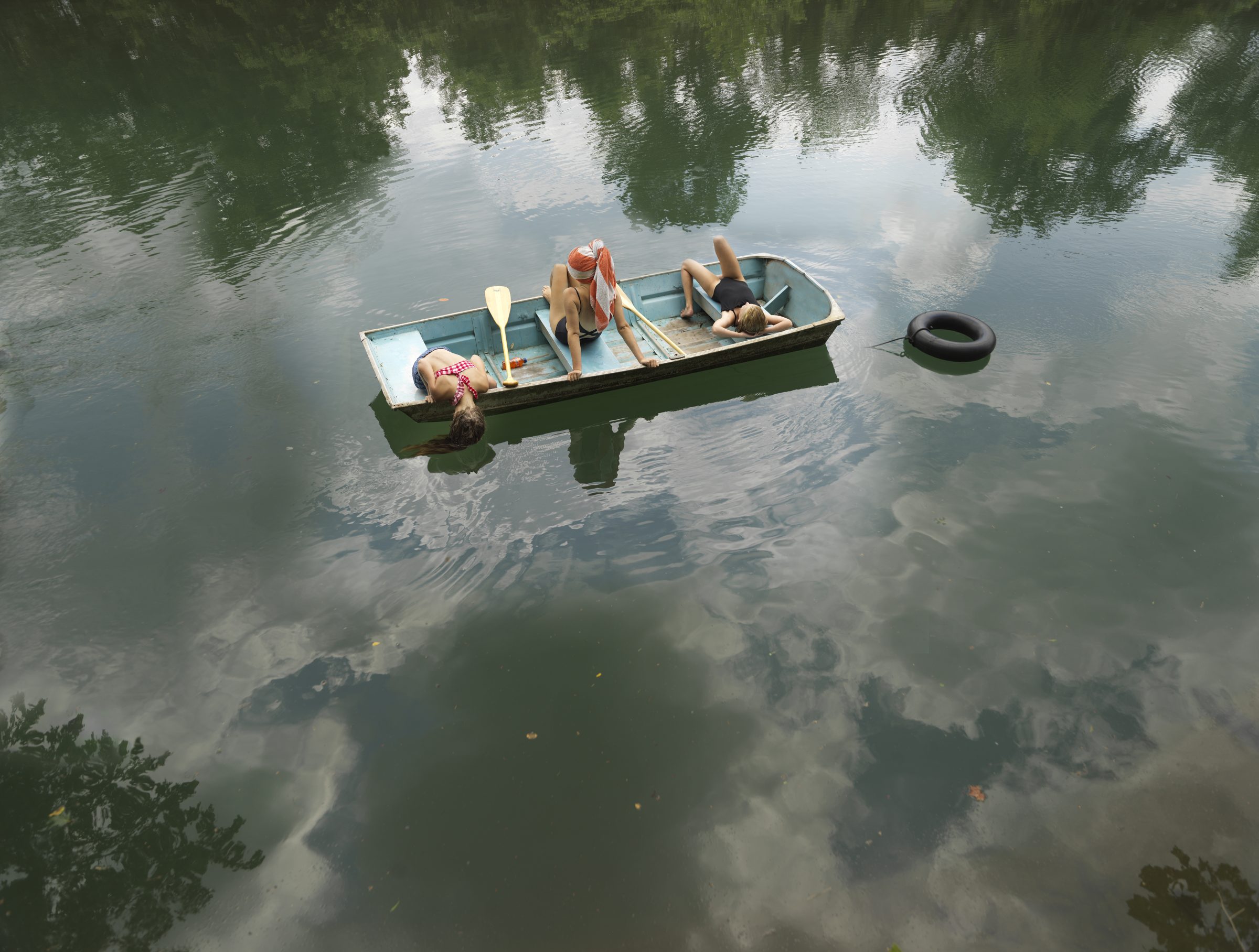 A small rowboat floats on still water surrounded by green trees and their reflections. Three figures recline in the boat amid oars and supplies, while a dark tire bobs nearby in the calm, mirror-like surface.