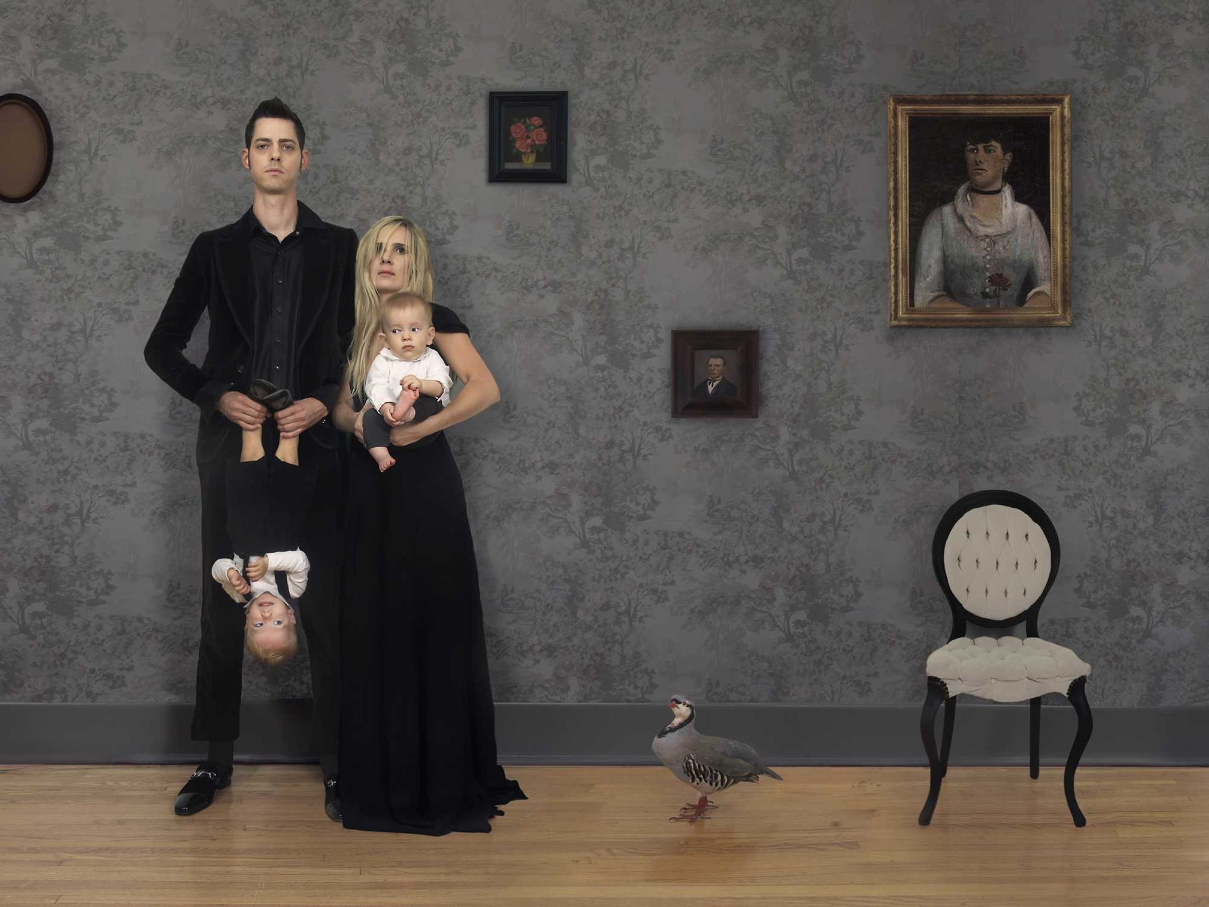 A man and woman in black clothing stand against a damask-patterned wall, with the woman holding two infants. A child hangs upside down from the man's arm, and a small goose walks across the wooden floor in the foreground. Period portraits and a round-backed chair are mounted on the wall behind them.