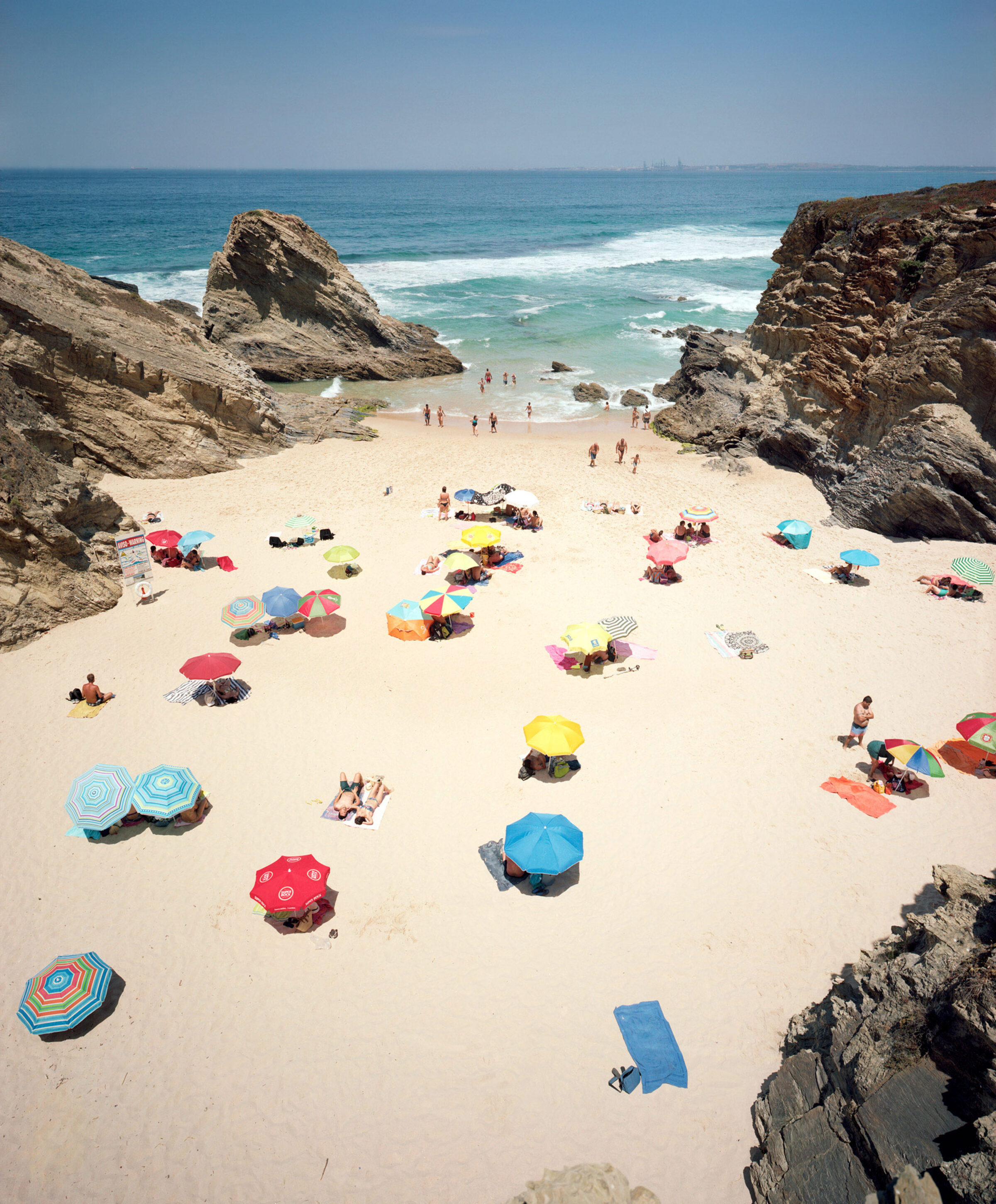 Aerial view of a sandy beach cove framed by dramatic rocky outcrops, dotted with colorful umbrellas in red, blue, yellow, and striped patterns. Beachgoers scatter across the pale sand while waves break along the shoreline, with a calm blue ocean and distant horizon visible beyond.