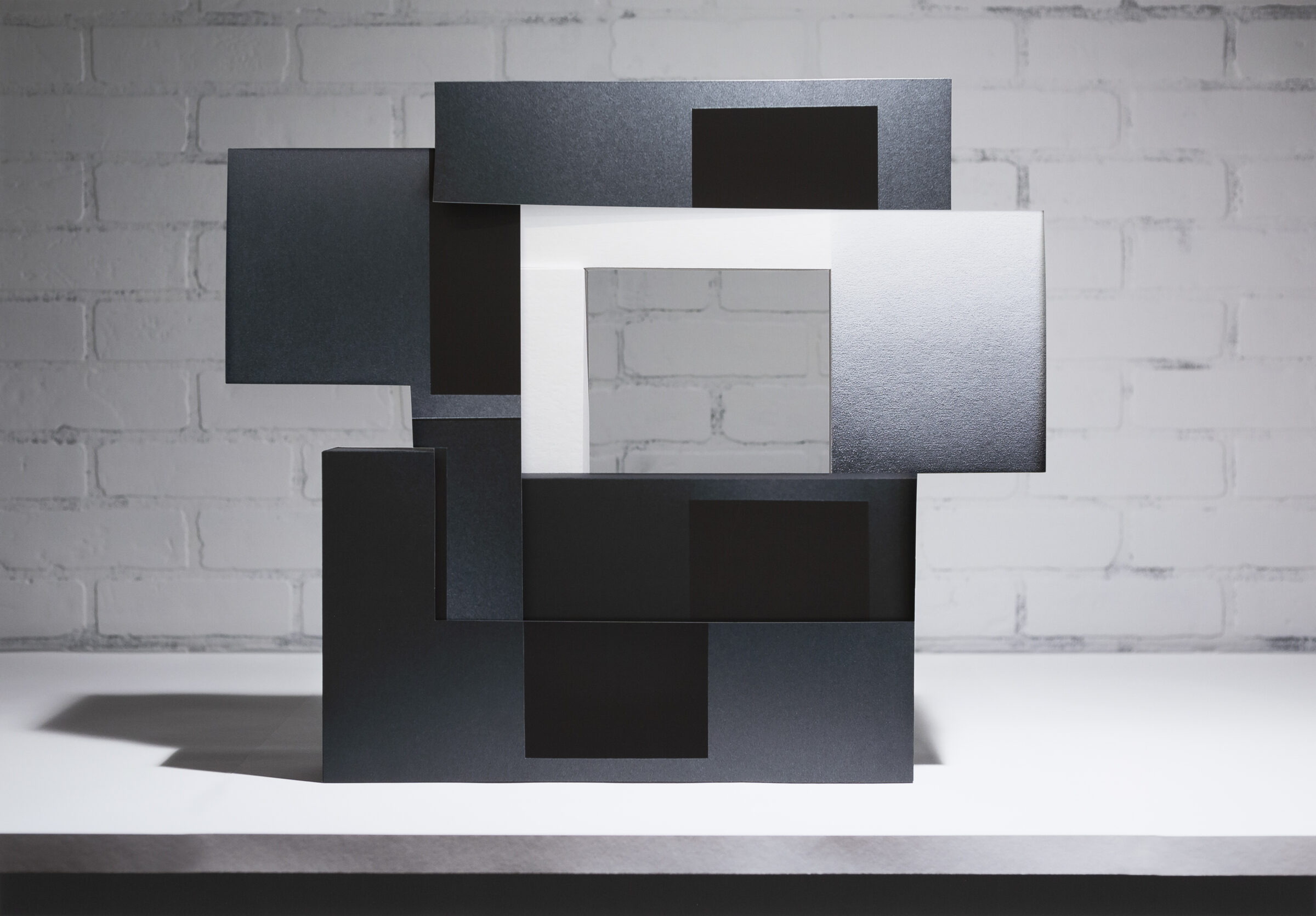 Layered rectangular panels in black, dark gray, and white arranged in an overlapping, three-dimensional composition against a white brick wall. The stacked forms create depth through varying planes and tones, with some surfaces appearing textured or printed, casting subtle shadows on the white surface beneath.
