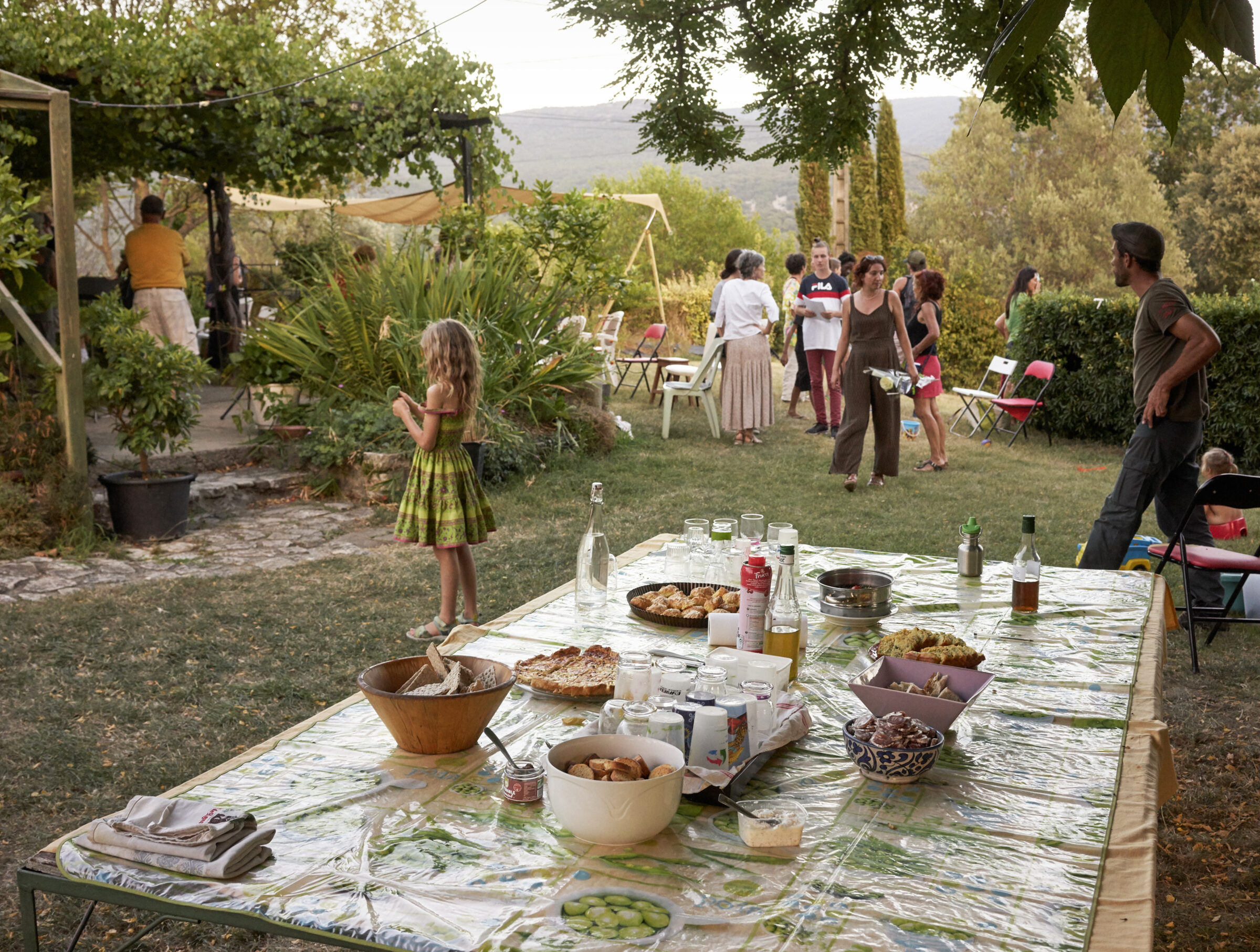 A garden gathering in a sunlit Tuscan-style landscape, with a long outdoor table laden with food and bottles in the foreground, and multiple guests mingling across a terraced lawn scattered with chairs and potted plants, mountains visible in the hazy distance.