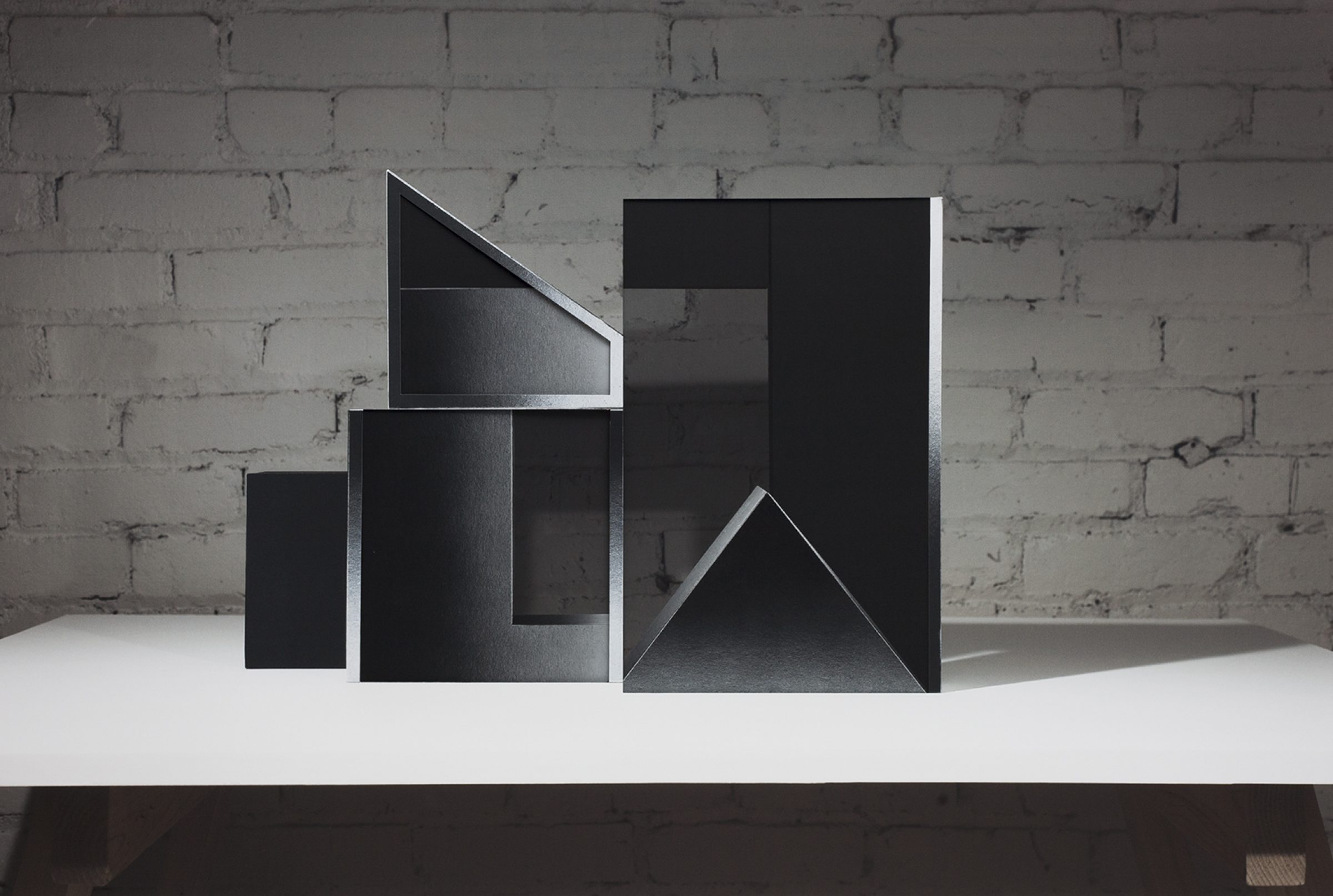 Abstract geometric sculpture made of black layered materials arranged in ascending rectangular and triangular forms on a white platform. The composition features sharp angles, cutouts, and overlapping planes that create dramatic shadows against a weathered brick wall.