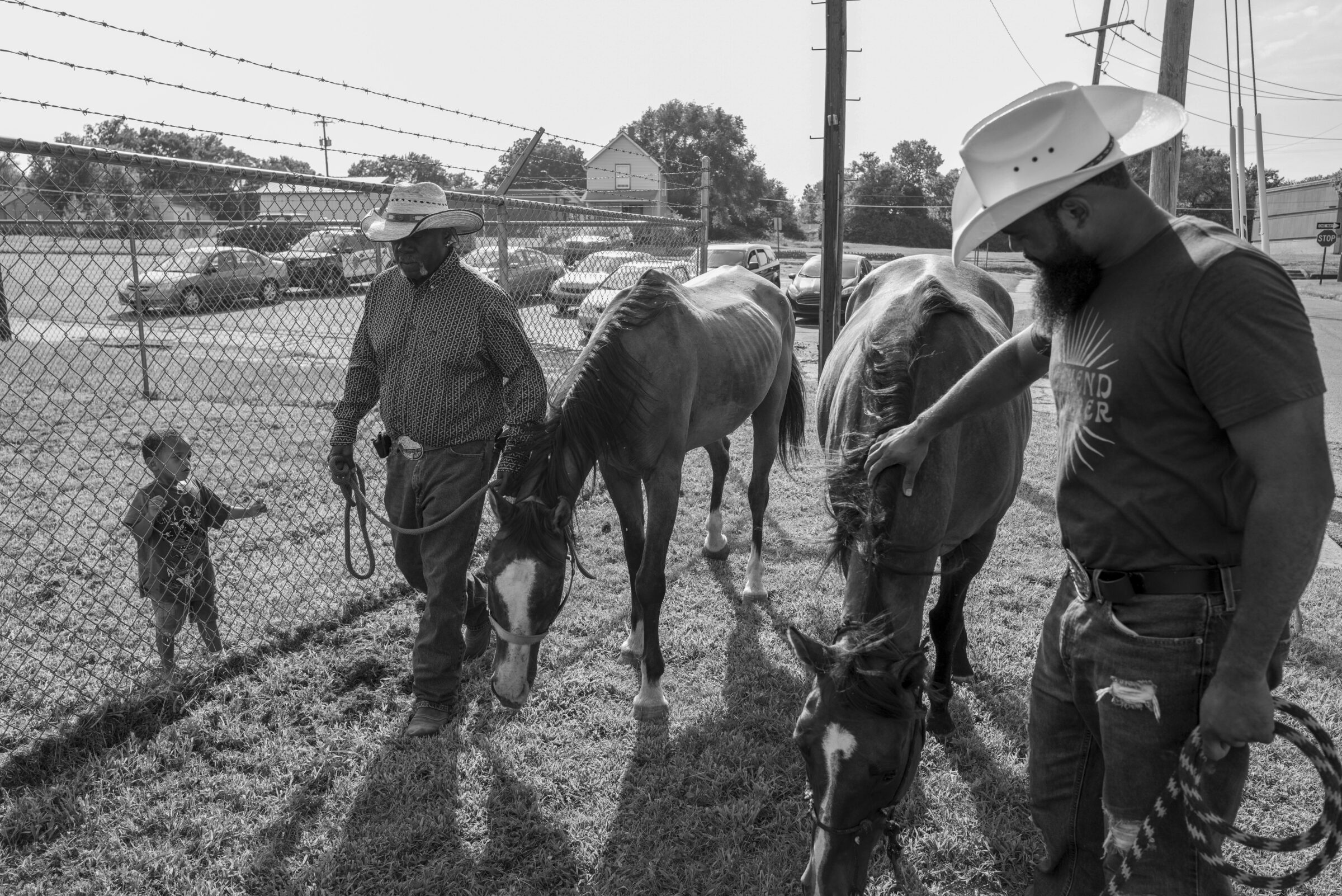 Black and white photograph showing two cowboys in cowboy hats leading horses through a ranch enclosure, with a small child reaching toward the fence in the foreground. Houses, utility poles, and chain-link fencing are visible in the background under an overcast sky.