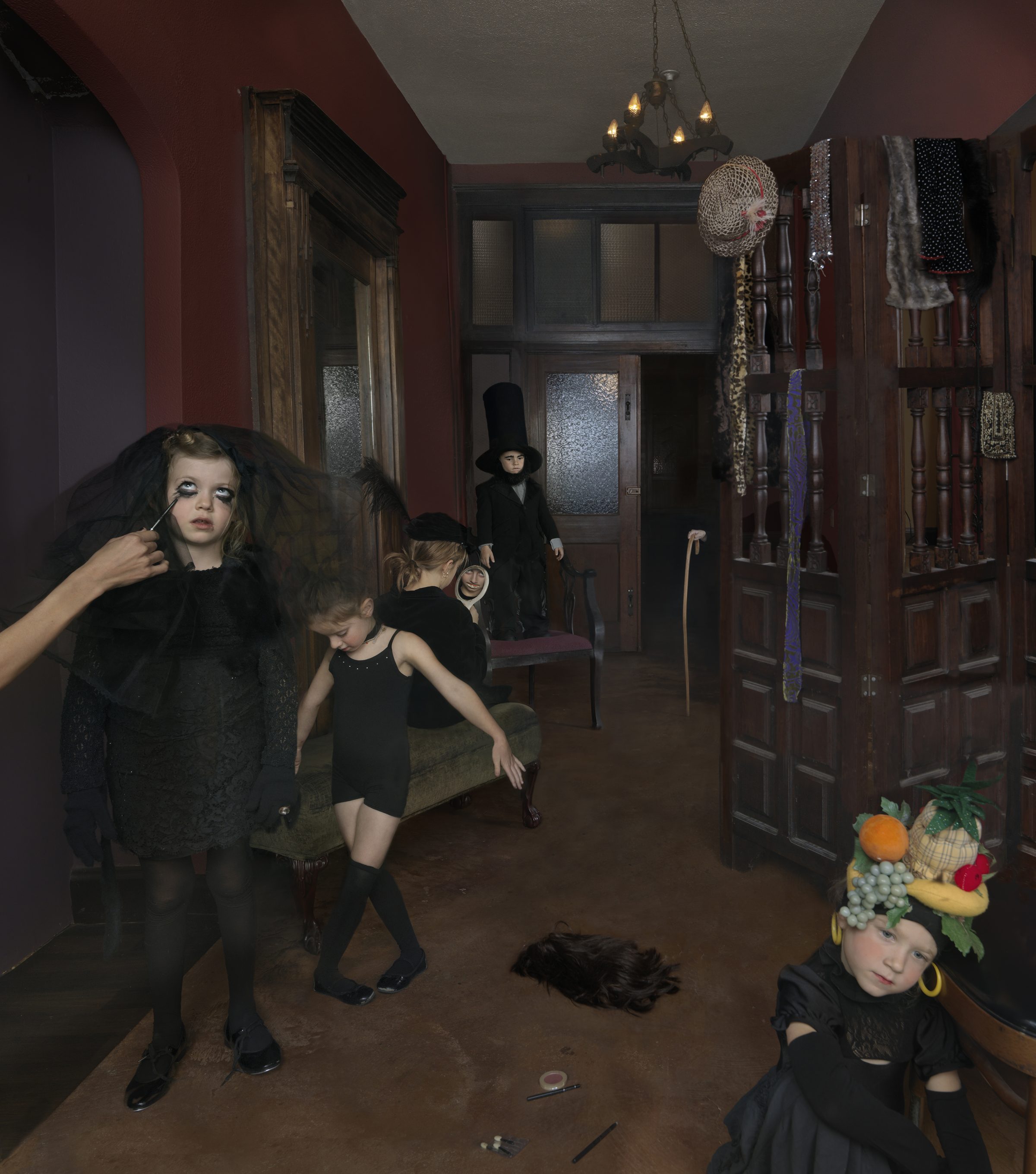 Children in dark clothing and theatrical makeup occupy a burgundy-walled room filled with wooden furniture and vintage accessories. A figure applies makeup to a child in the foreground while others pose throughout the space, one wearing an elaborate fruit and vegetable headdress, in a staged domestic scene with surreal, darkly whimsical undertones.