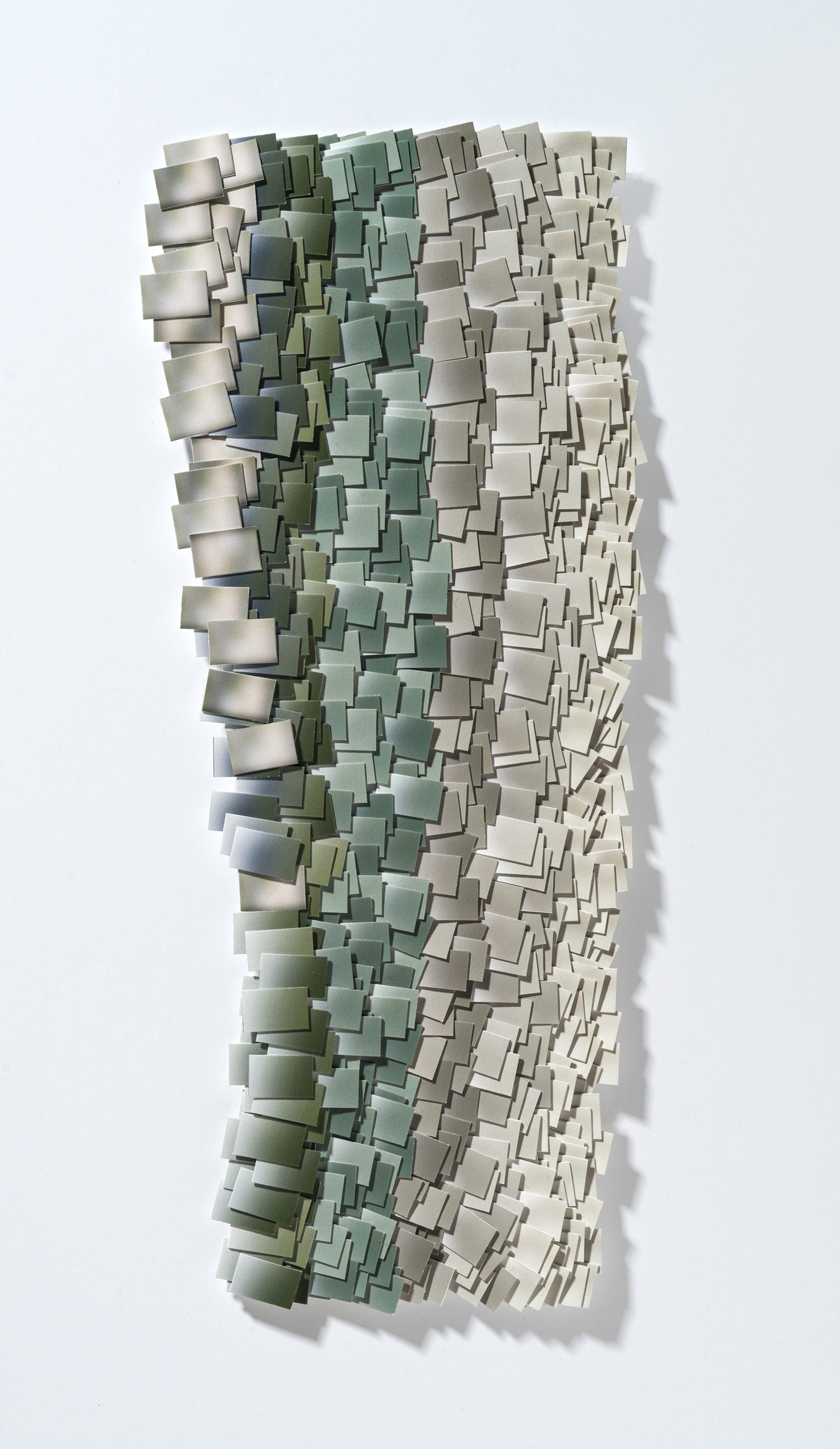 A vertical wall-mounted artwork composed of hundreds of hand-cut and layered rectangular paper fragments in graduated tones of sage green, taupe, and cream, creating a three-dimensional textured surface with pronounced shadows that suggest a pixelated or topographic landscape.
