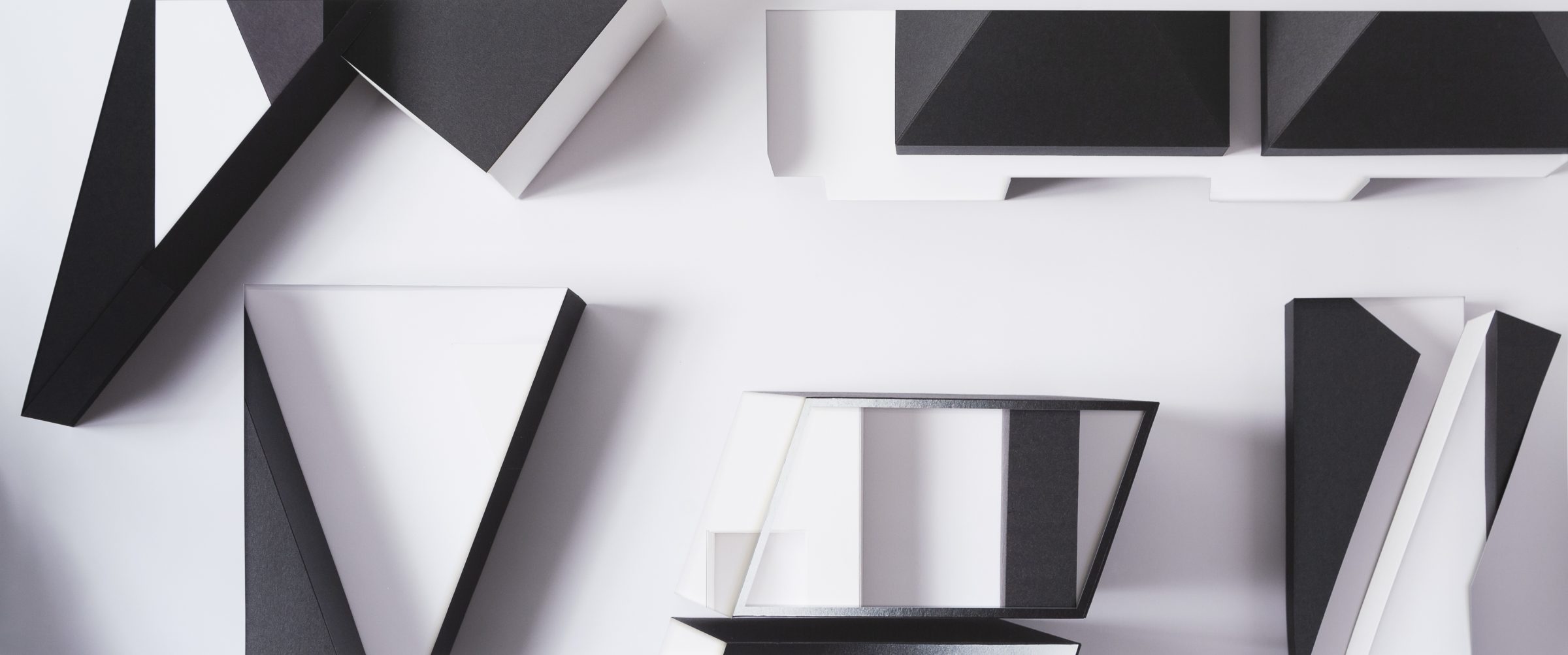 Geometric abstract composition of layered, fragmented forms in black, white, and gray tones arranged across a light background. Sharp angular shapes and rectangular planes overlap and cast shadows, creating depth and visual tension through a deconstructed, collage-like arrangement.