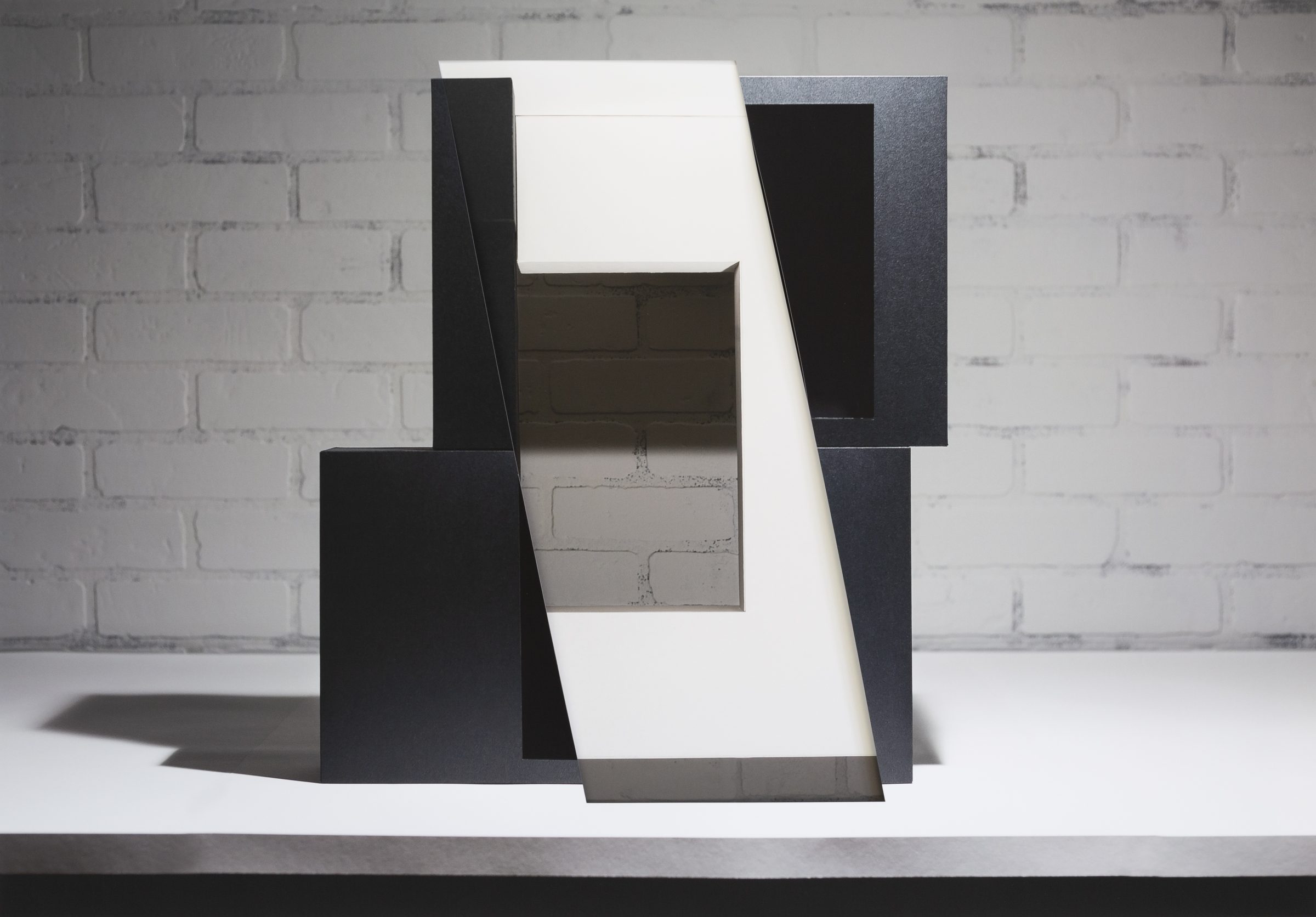 Abstract geometric composition featuring layered rectangular forms in black, white, and gray tones arranged to create depth and dimensional space. The collaged and layered construction reveals cut and overlapping planes that cast shadows against a white brick wall backdrop.