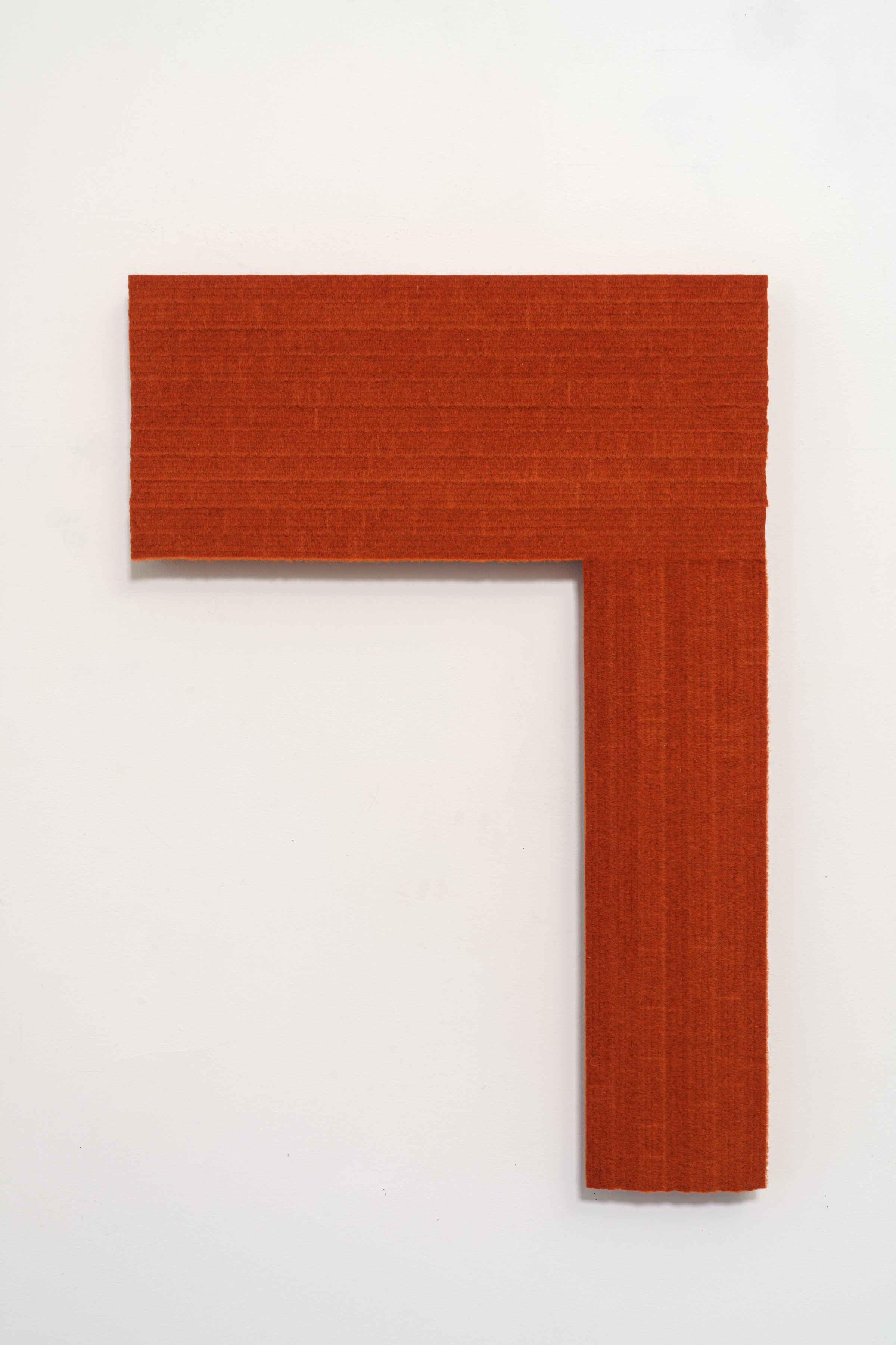 A bold rust-orange geometric form in the shape of a right angle or inverted "L" mounted on a white wall. The piece has a textured, matte fabric surface with subtle color variation.