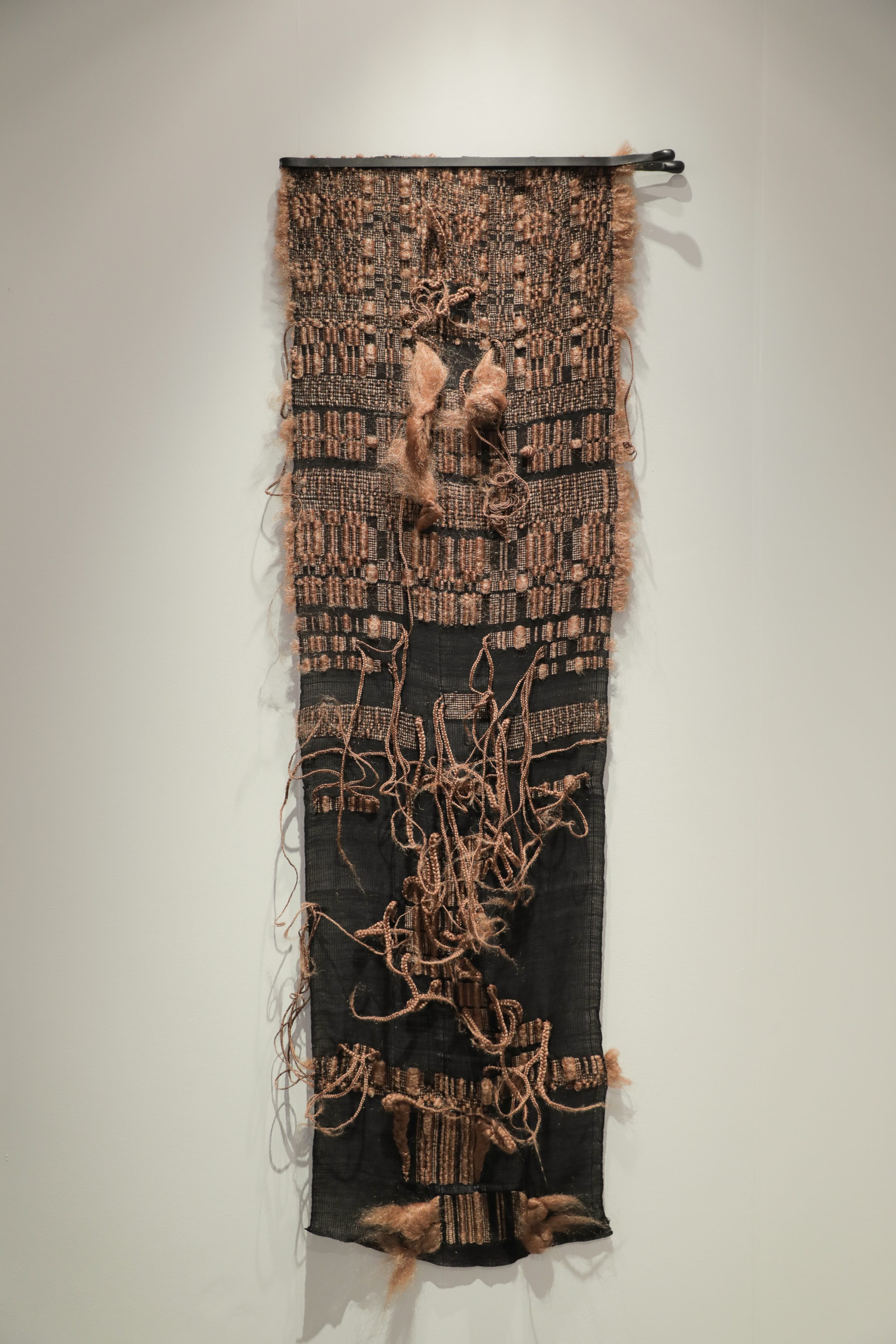 A vertical textile wall hanging combining woven cotton with synthetic hair elements. The upper portion features a tan and black woven pattern with figurative forms, while the lower section is predominantly black with looping strands of blonde hair creating organic, gestural marks across the surface.