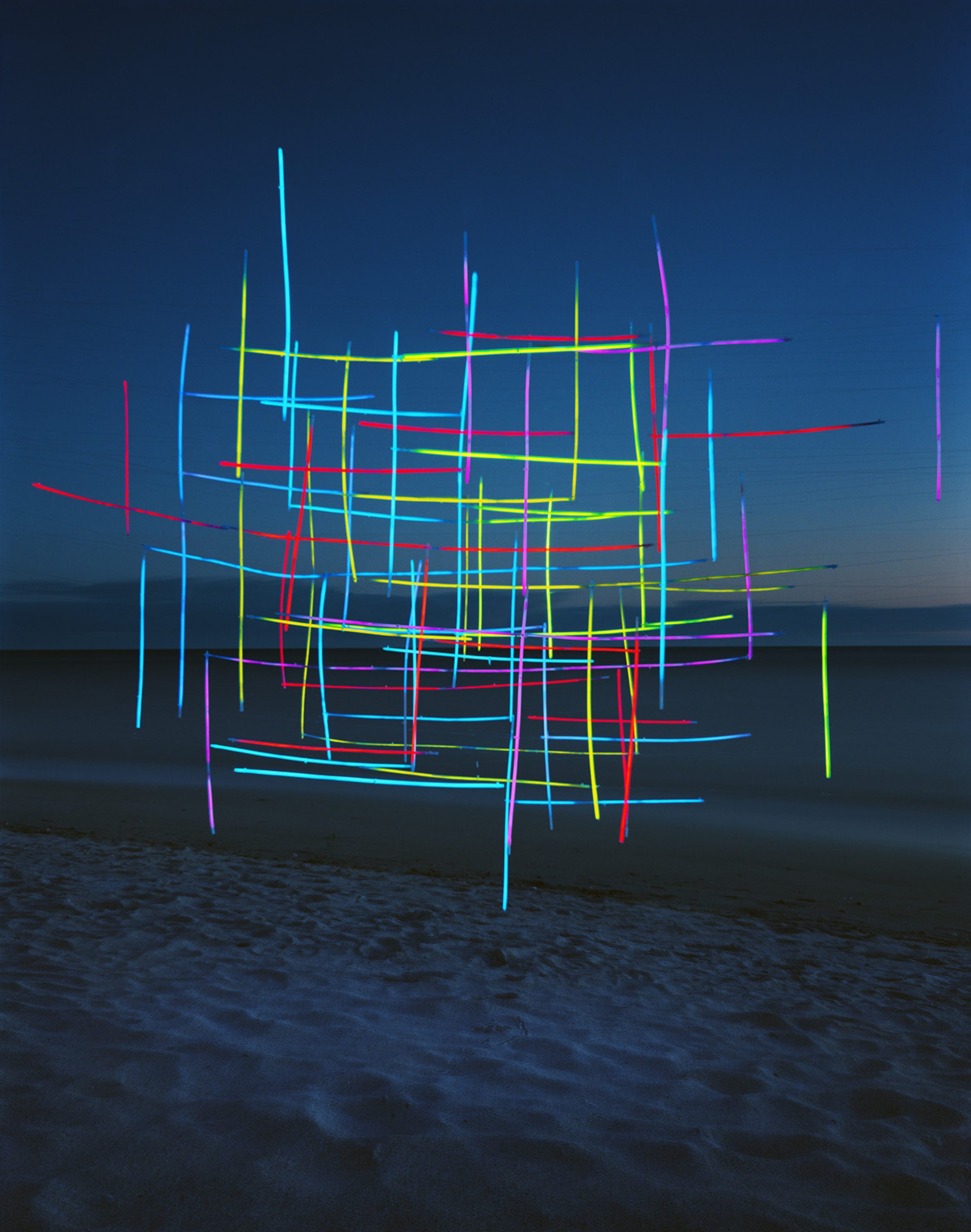 Neon-colored lines in cyan, yellow, red, magenta, and green form an intricate geometric lattice structure suspended on a beach at dusk, with the ocean and darkening sky visible behind it.