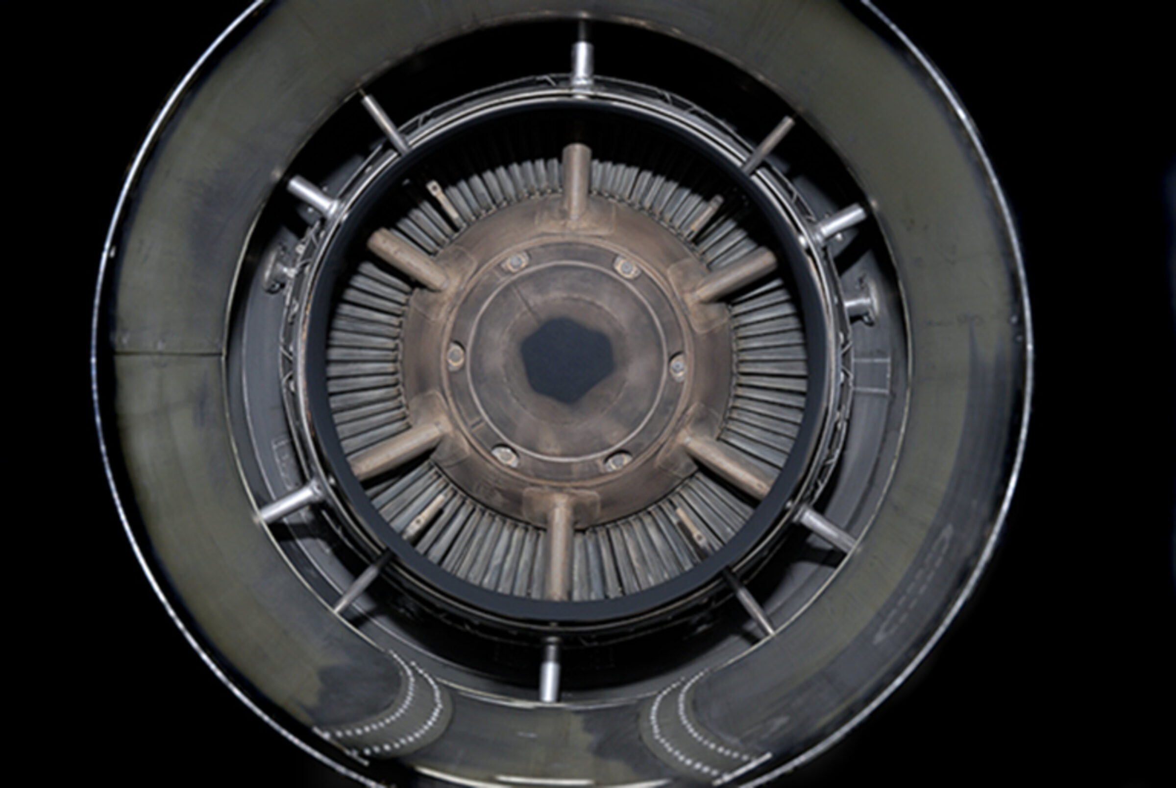 Concentric circular view of a mechanical turbine or engine intake, rendered in grayscale. Radiating fins and metallic components create a hypnotic, symmetrical pattern receding toward a dark central aperture, photographed directly from above against a black background.