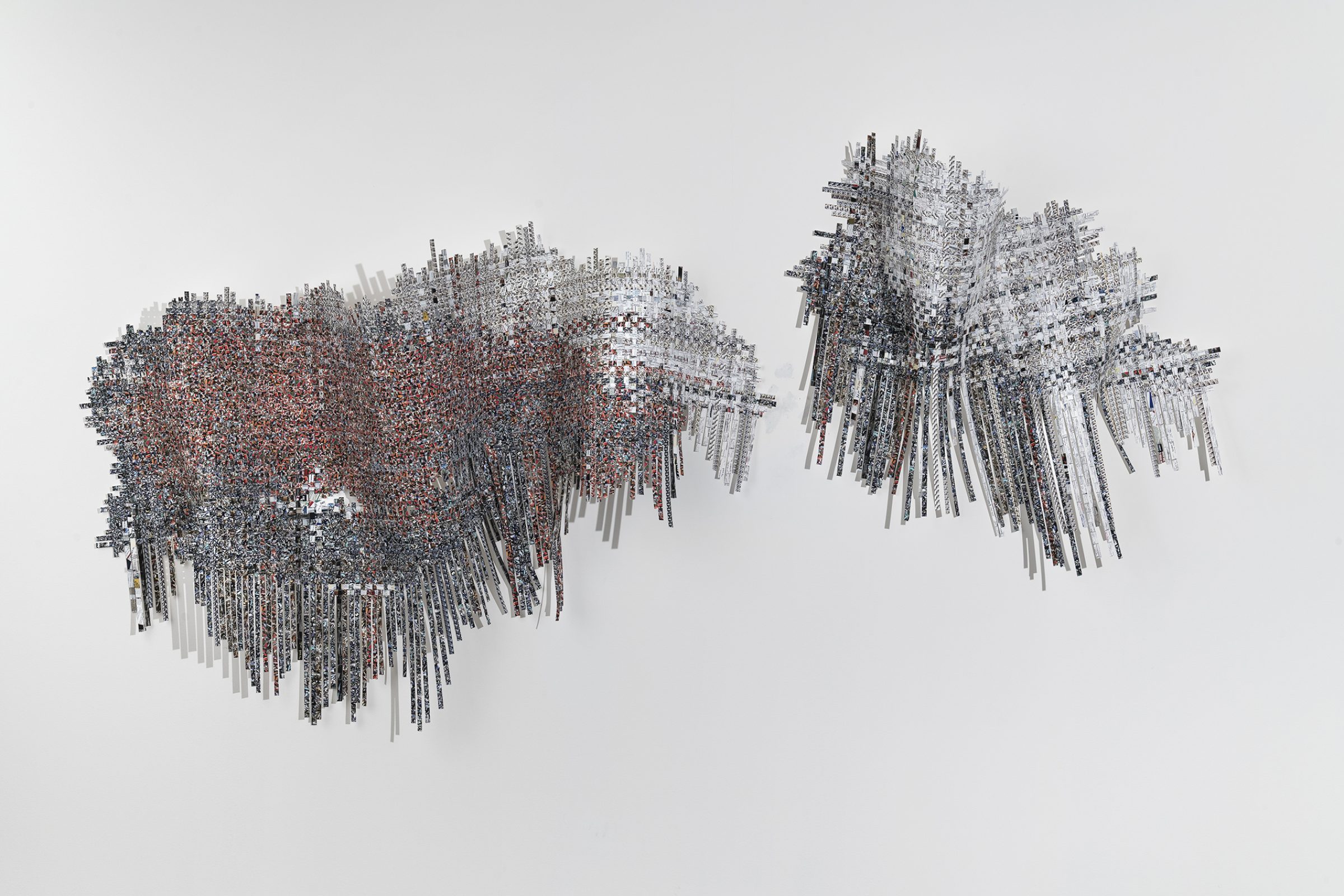 Two abstract sculptural forms composed of densely layered rectangular strips and particles suspended in space against a neutral background. The left form features warm reddish-brown and gray tones with a more compact, cloud-like density, while the right form displays predominantly gray and white tones in a more dispersed, wispy configuration. Both structures create depth through their three-dimensional arrangement of fragmented materials.