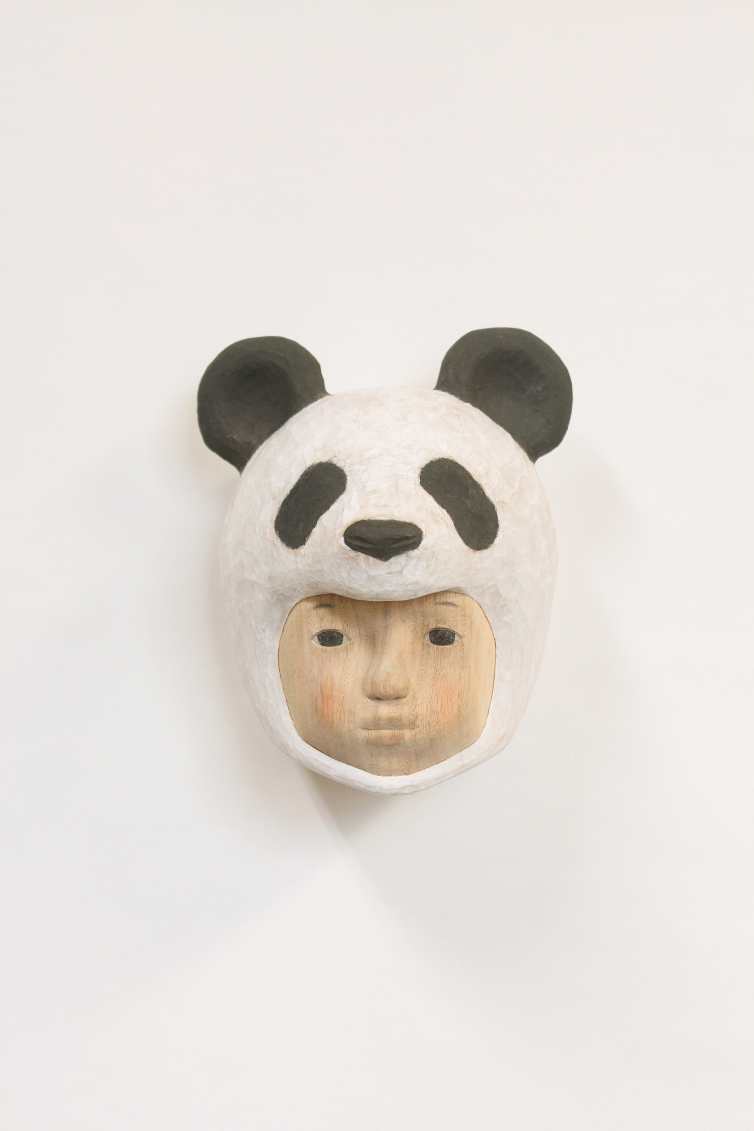 A carved wooden mask depicting a human face wearing a giant panda head covering, with black and white coloring and subtle flesh-toned facial features below. The sculpture combines naturalistic human portraiture with stylized animal characteristics, mounted on a white background.