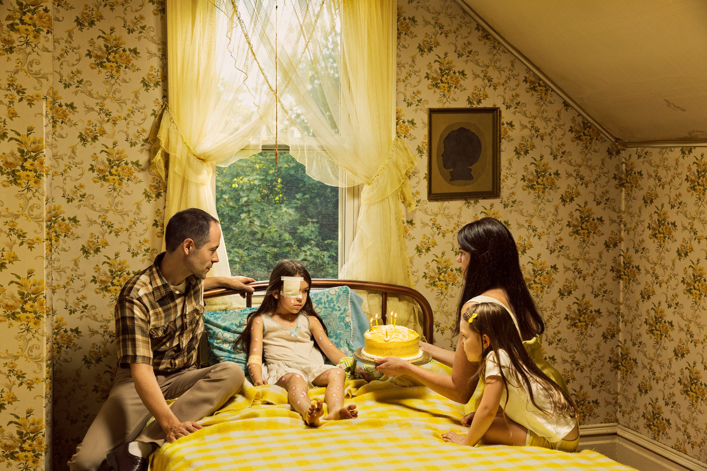 A family of four sits on a yellow striped bed in an attic room with floral wallpaper and yellow curtains framing a window overlooking green foliage. A yellow birthday cake with lit candles sits on the bed as the father touches his young daughter's head while the mother and older daughter look on.