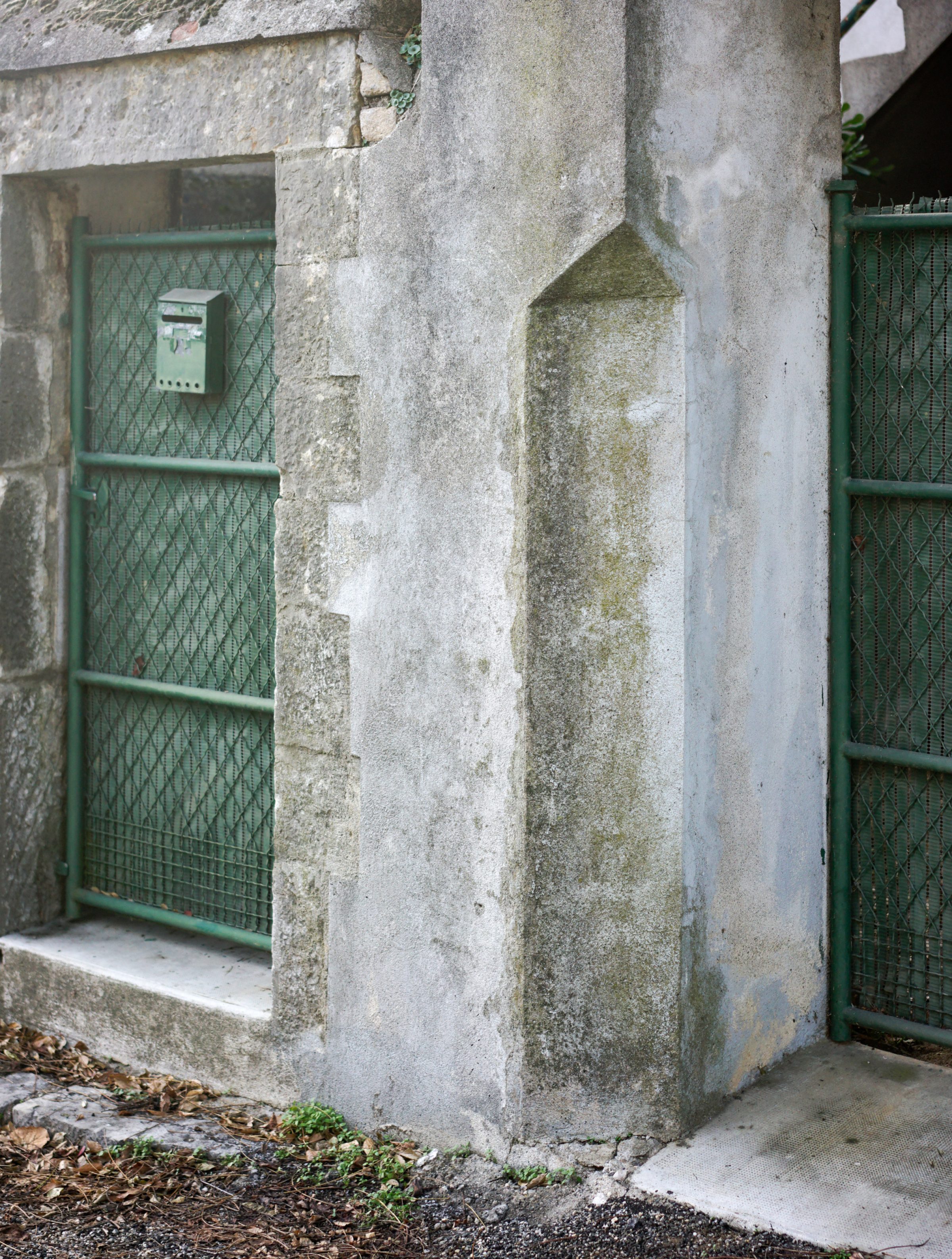 Weathered concrete pillars and walls marked by moss and lichen staining frame a narrow passageway lined with green metal gates and a mailbox, capturing the texture and decay of an industrial or utilitarian structure.
