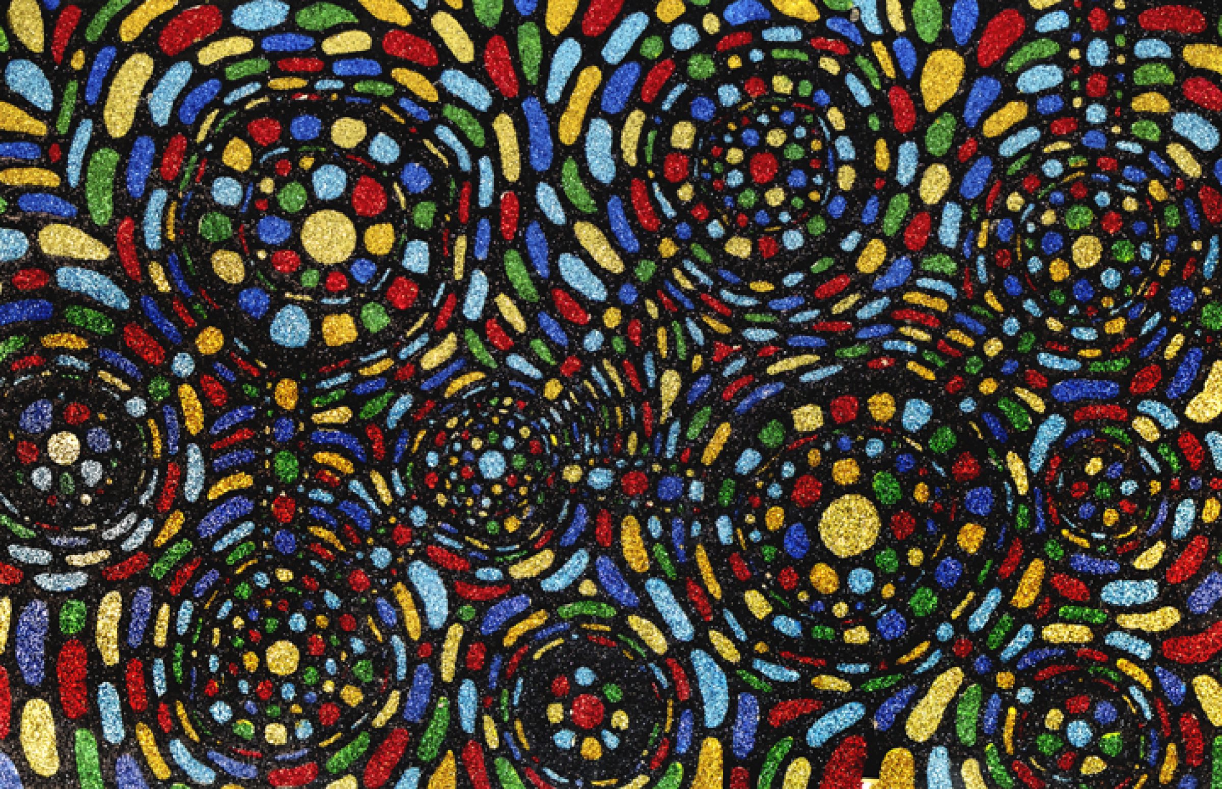 Vibrant abstract composition featuring concentric circular swirls rendered in bold primary colors—red, blue, yellow, and green—with shimmering glitter and textured cut paper creating dynamic movement across the dark background.