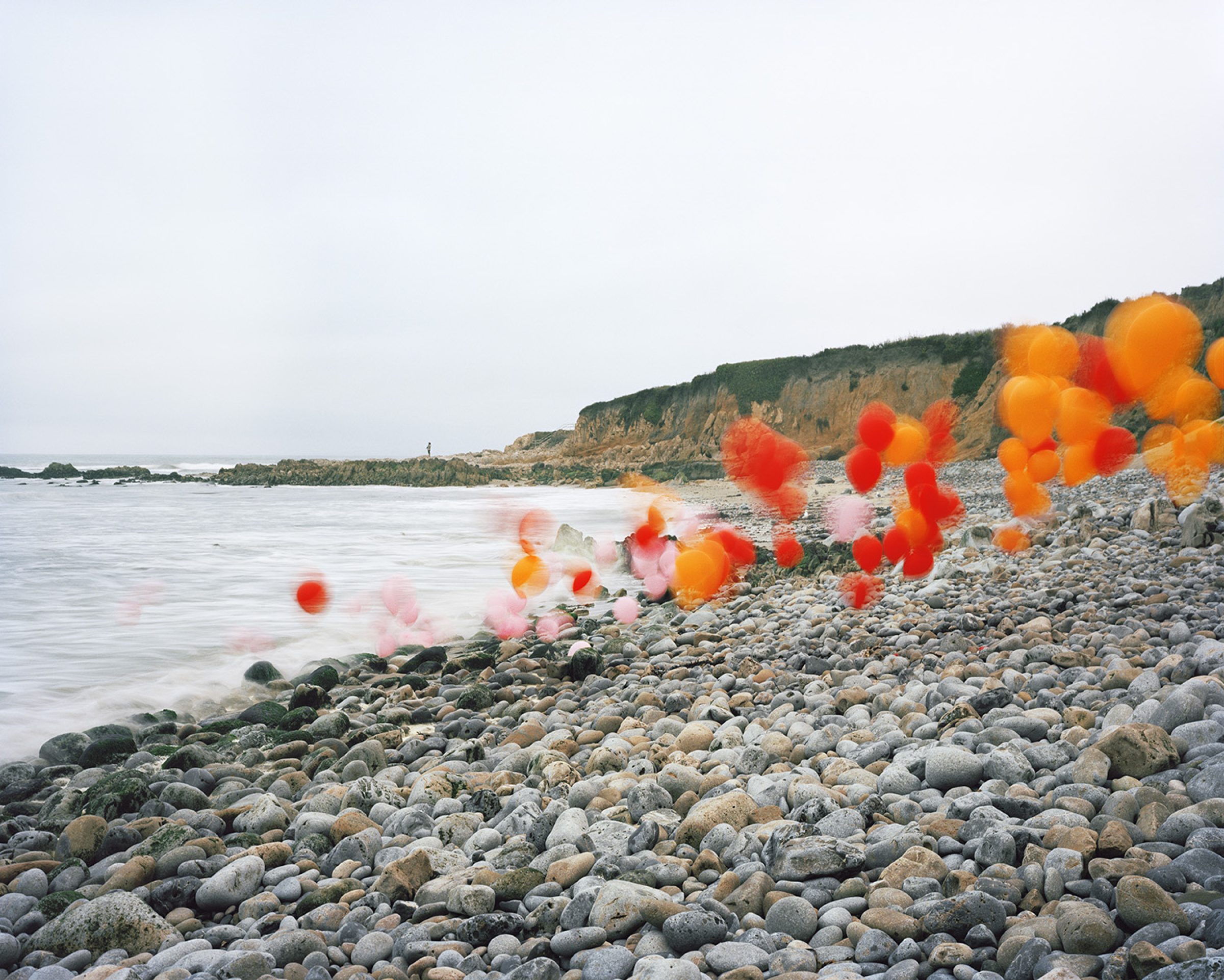 Vibrant red and orange balloons float and cluster along a rocky beach, some blurred in motion, creating a striking contrast against the neutral tones of rounded stones, calm water, and distant cliffs under an overcast sky.