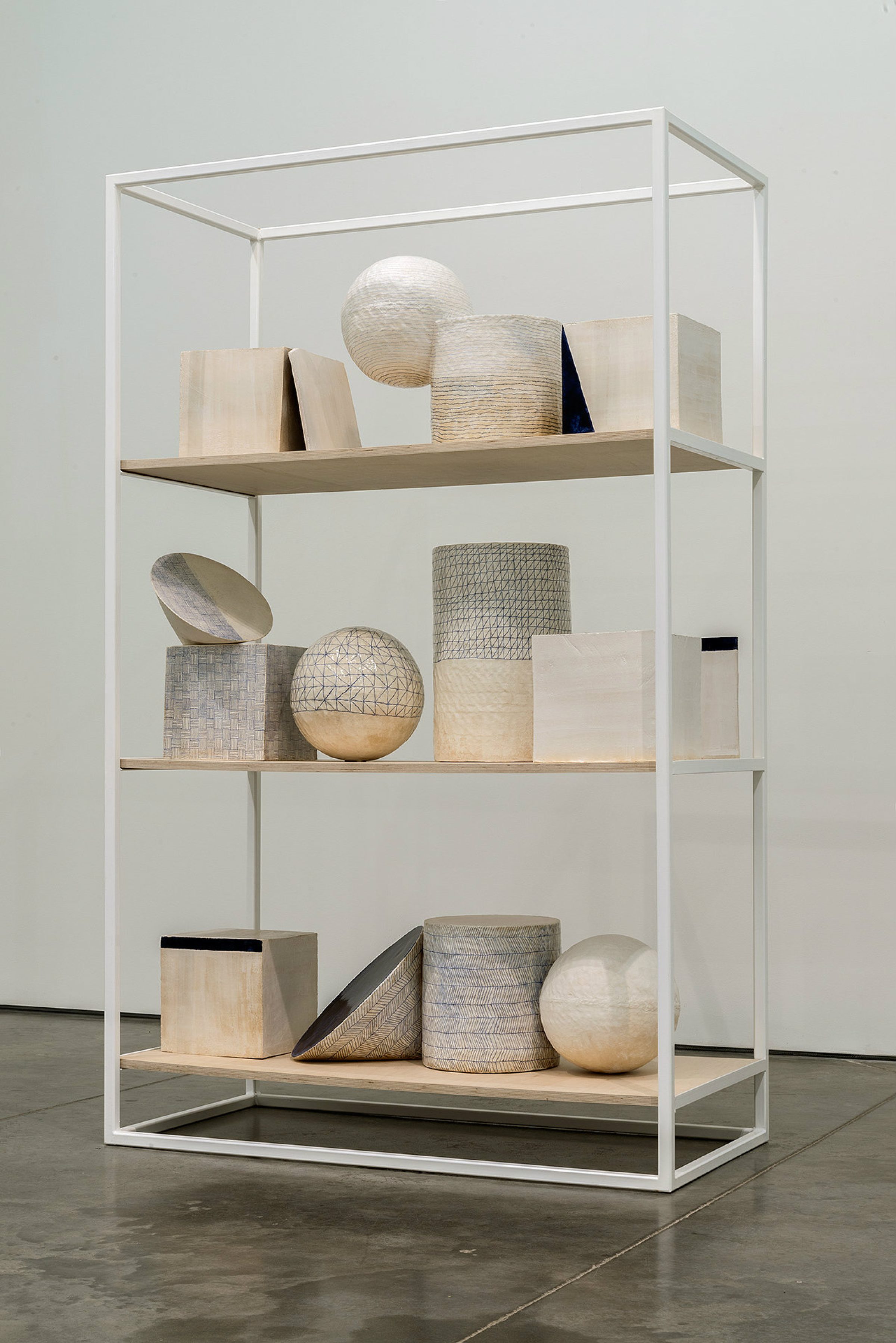 White steel display case with four wooden shelves containing an arrangement of geometric sculptural objects in natural materials including woven baskets, spheres, cylinders, and rectangular forms in cream, beige, gray, and black tones.