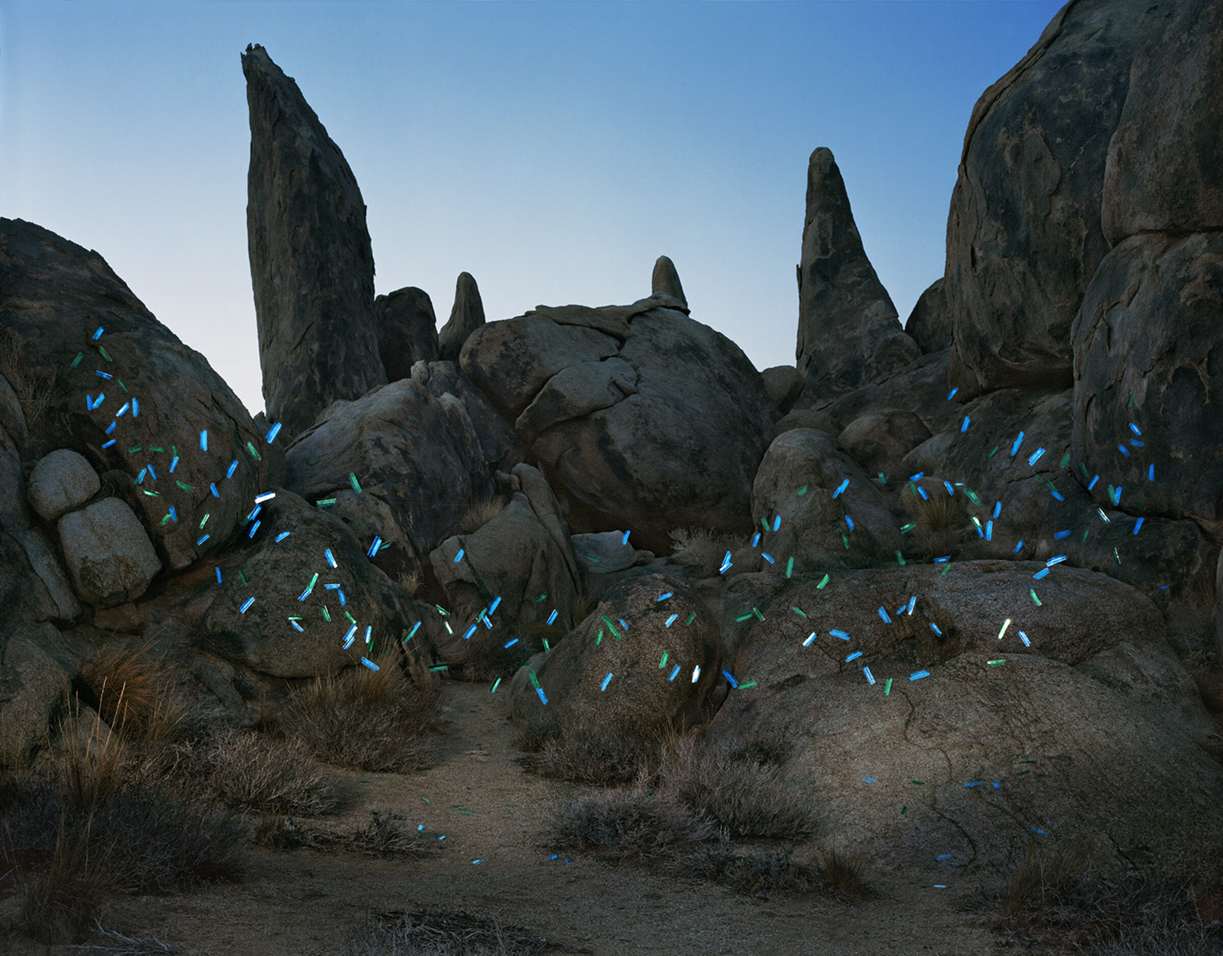 Desert rock formation with tall granite boulders and pinnacles against a clear blue sky, overlaid with bright blue and green painted or digital file tab shapes scattered across the sandy ground and lower rock surfaces.