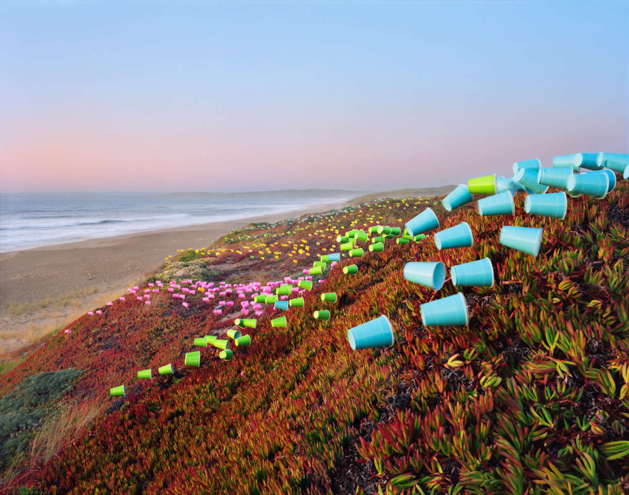 Colorful plastic cups arranged in geometric patterns across a coastal hillside covered in succulent vegetation, with a beach and ocean visible in the background at dusk.