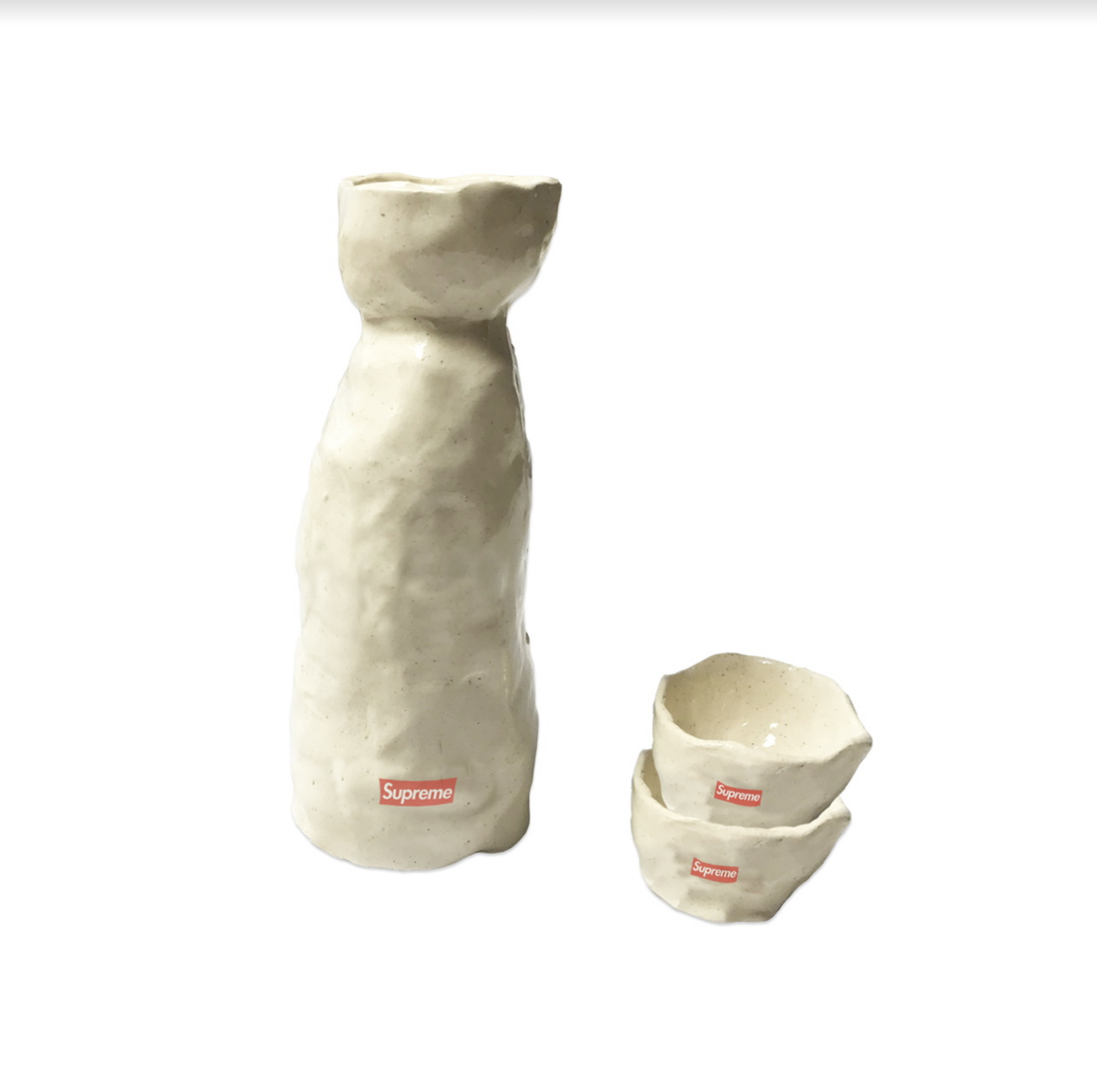 Cream-colored ceramic sake set consisting of a tall bottle with a tapered cylindrical body and flared rim, alongside two stacked small bowls with organic, slightly irregular edges. Small red rectangular logos appear on each piece against the pale, speckled glaze surface.