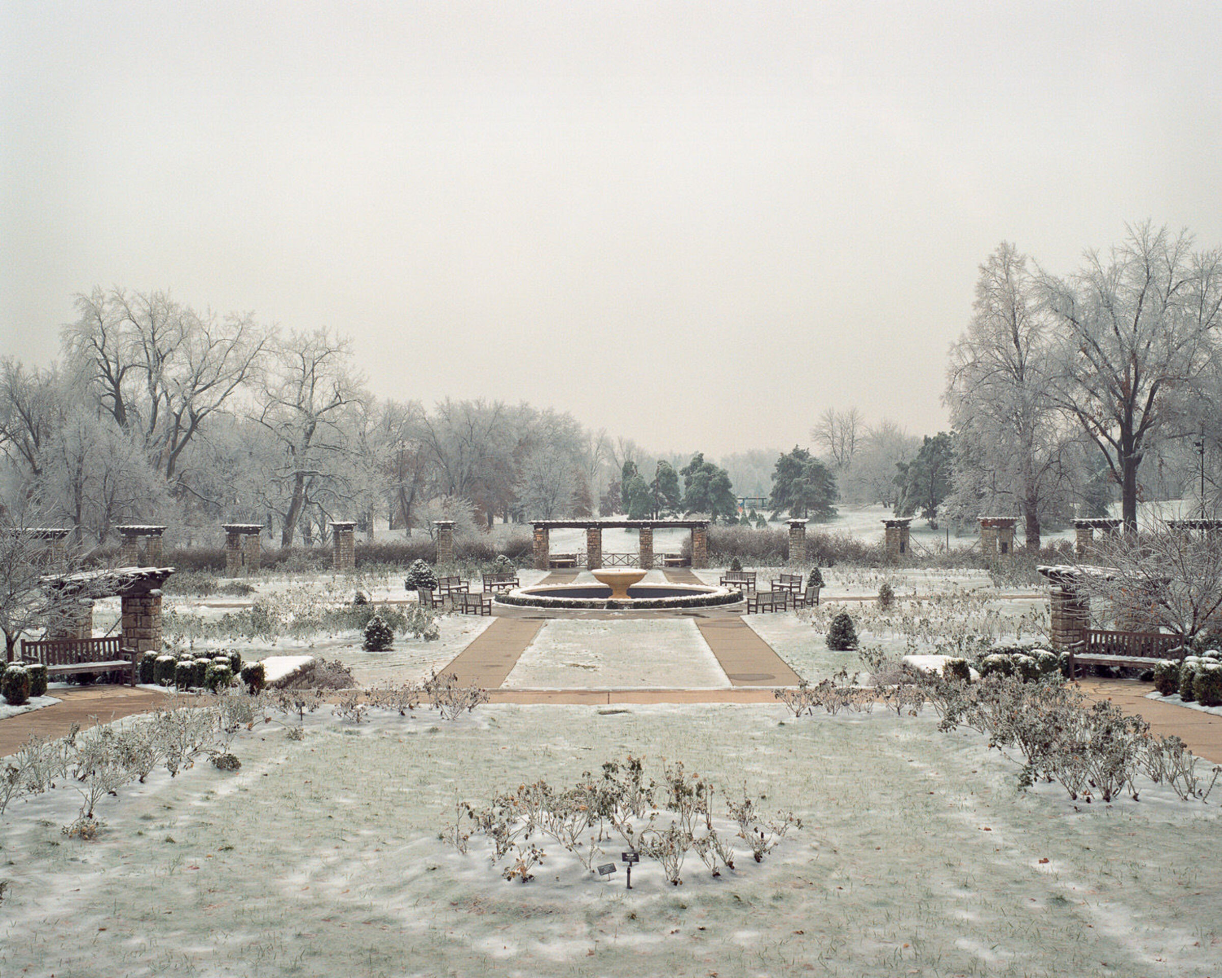 A frost-covered formal garden with a central circular fountain viewed down a symmetrical pathway. Bare trees frame the scene on both sides while brick structures and evergreens line the composition, all blanketed in ice and snow beneath an overcast sky.