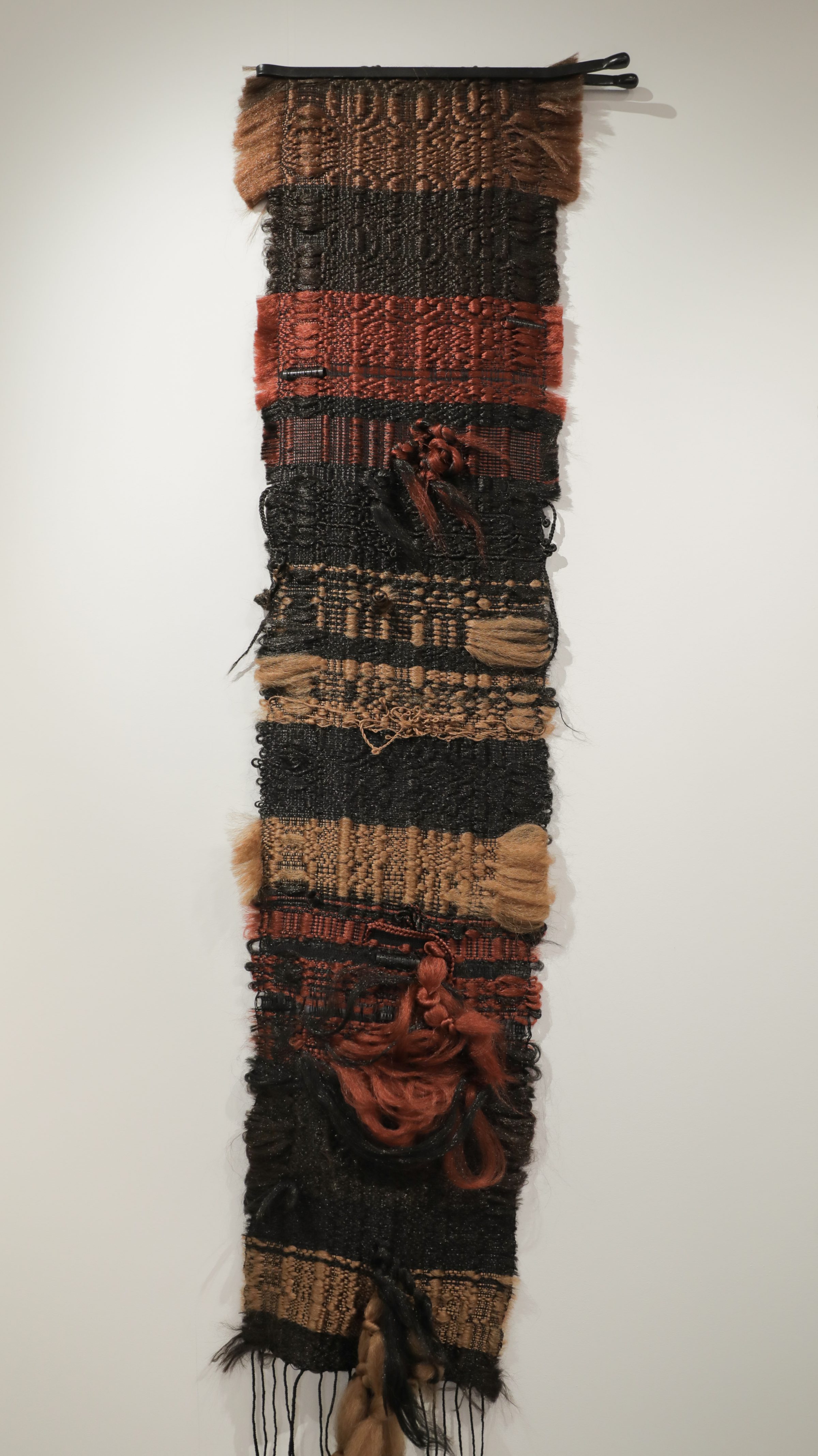 A vertical textile artwork composed of stacked fabric strips in varying weaves and textures, featuring earthy tones of brown, black, rust red, and tan. The composition includes woven sections, synthetic hair elements, and sculptural components including plastic rollers and bobby pins integrated throughout, with loose fringe at the bottom.