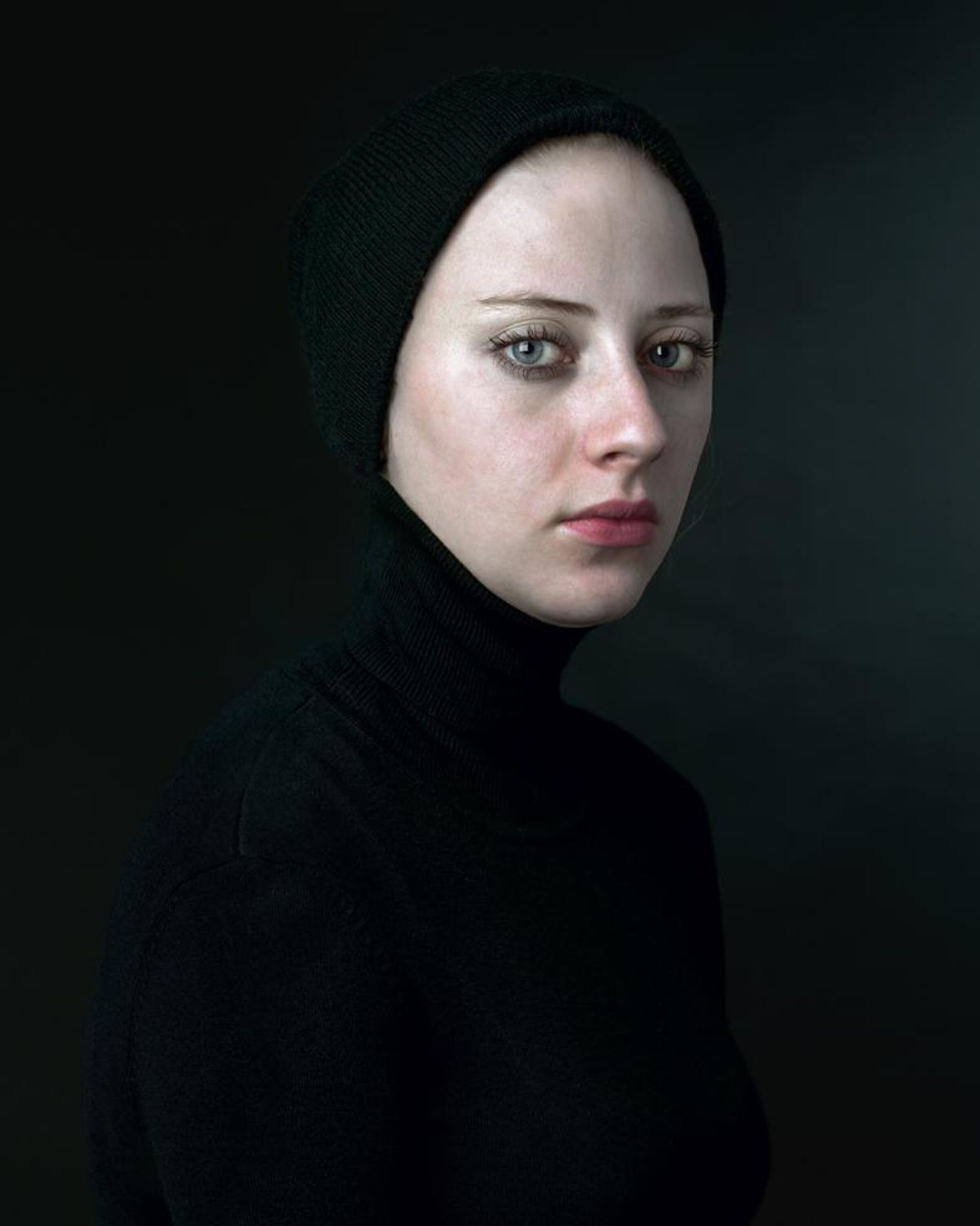 Portrait of a pale-skinned person wearing a black knit hood and turtleneck, photographed against a dark background in three-quarter view. The subject gazes downward with a contemplative expression, their face luminously lit against the shadowed composition.