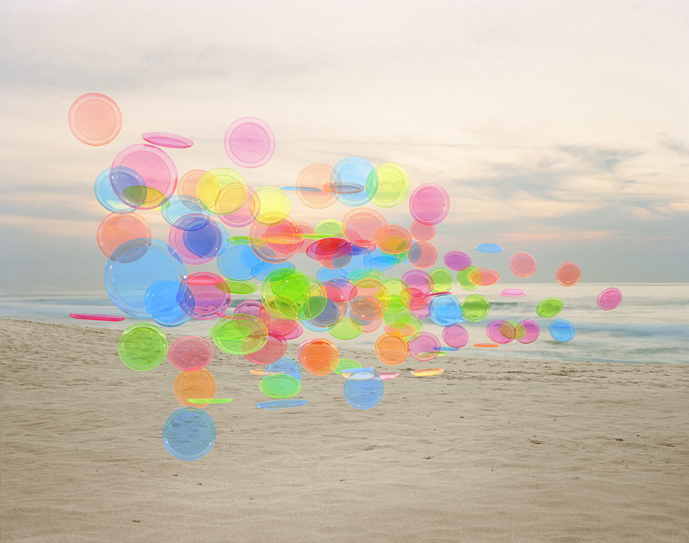 Translucent colored plastic discs and utensils in vibrant hues of blue, pink, green, yellow, and orange are arranged on a sandy beach, creating an abstract composition against a calm ocean and overcast sky.