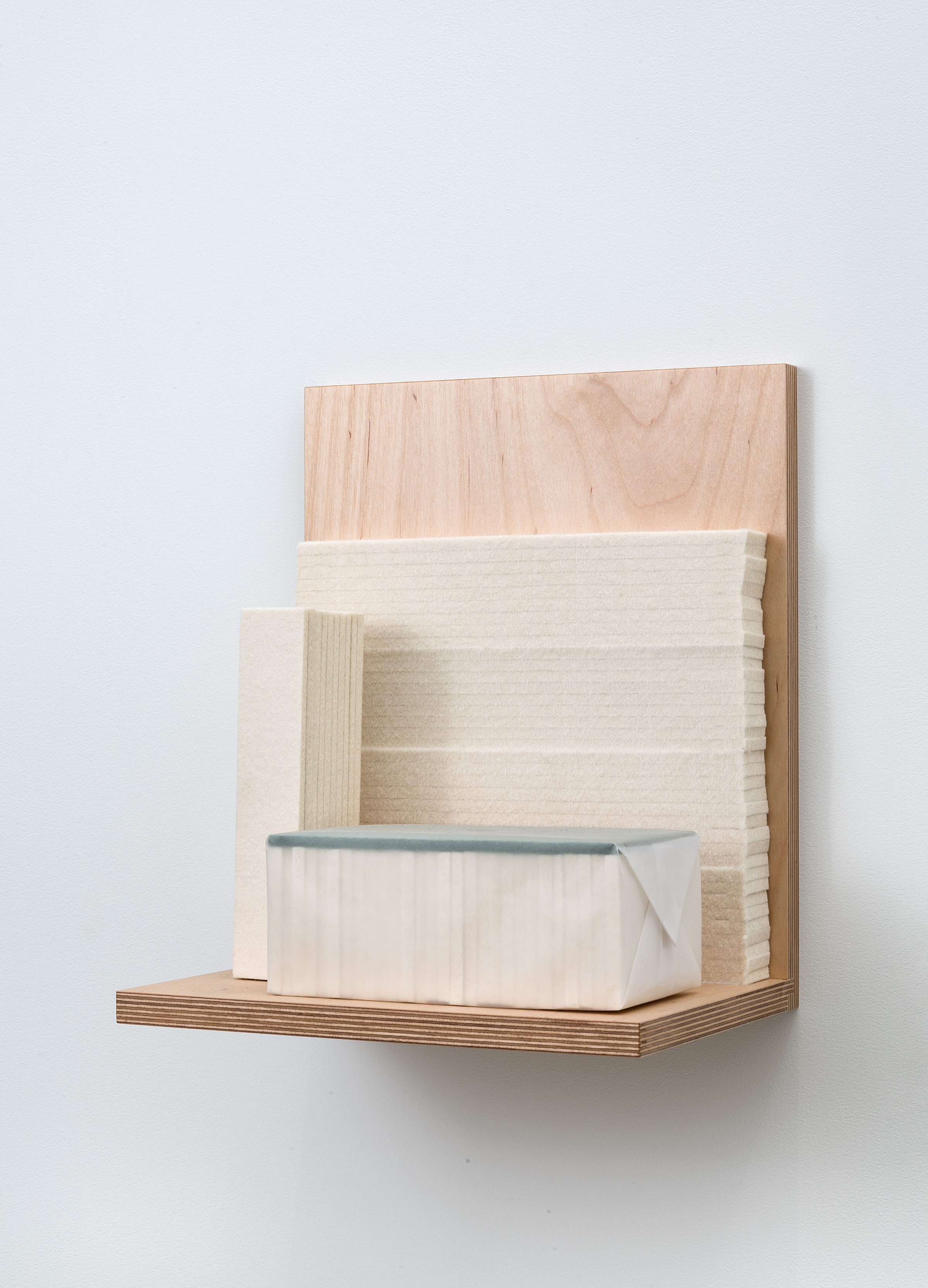 A wooden wall-mounted shelf unit constructed from light plywood and natural wood veneer, featuring geometric compartments and a cream-colored fabric-wrapped box with a blue-gray felt edge resting on the lower shelf.