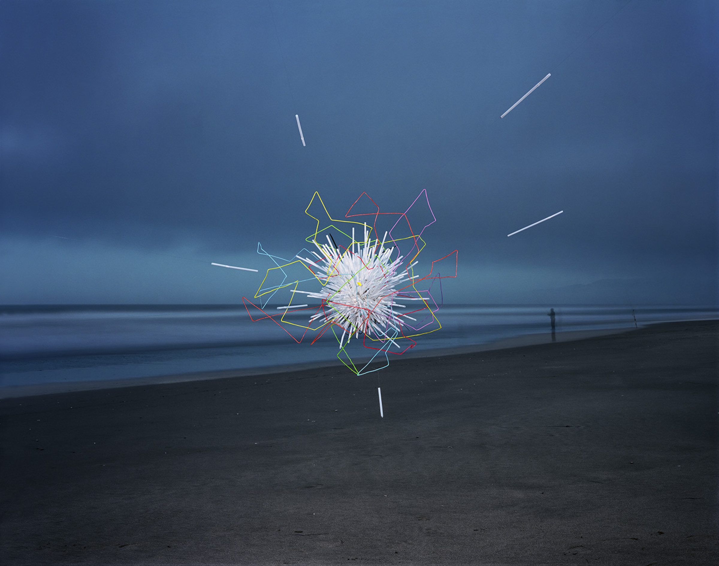 Neon-outlined geometric shapes in yellow, red, pink, and white radiate outward from a bright central burst on a dark beach, with horizontal straws extending across the composition against a moody blue-grey ocean and sky.