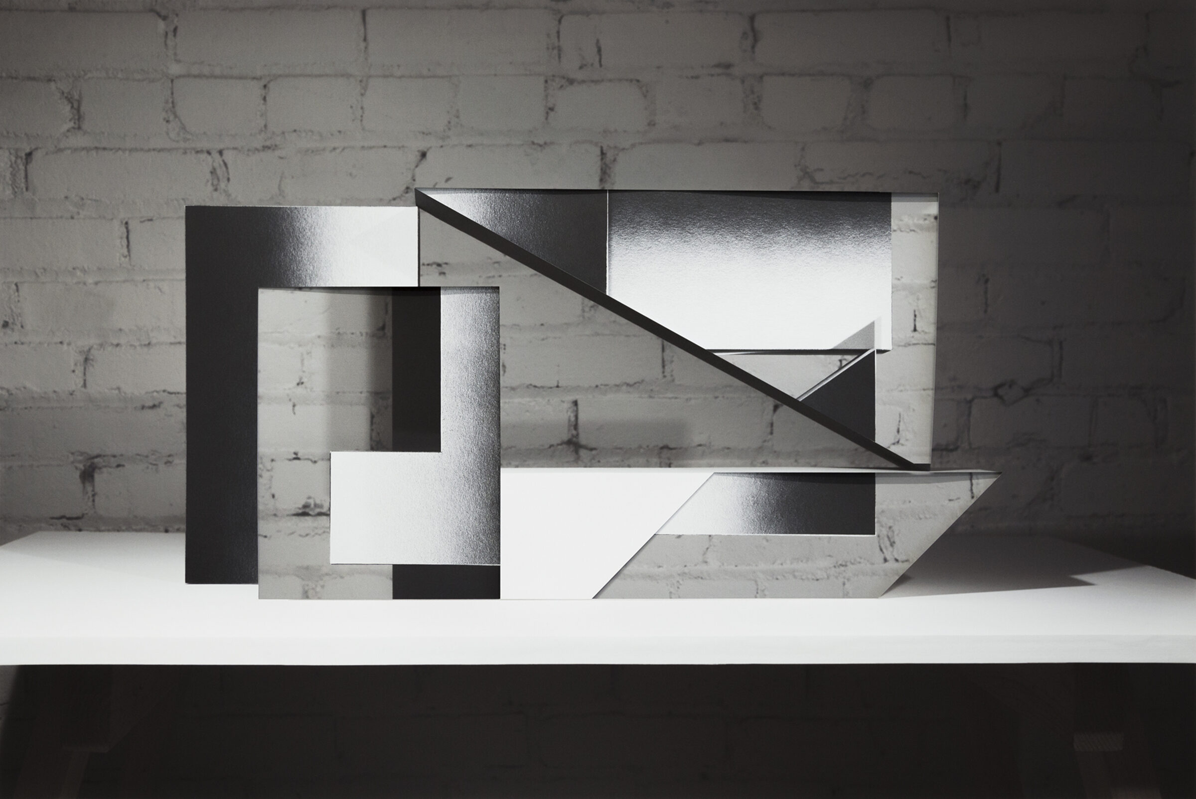 An abstract sculptural composition of layered black and white rectangular forms arranged on a white platform against a white brick wall. The overlapping planes create sharp angles and diagonal lines, with contrasting matte and reflective surfaces that cast distinct shadows.