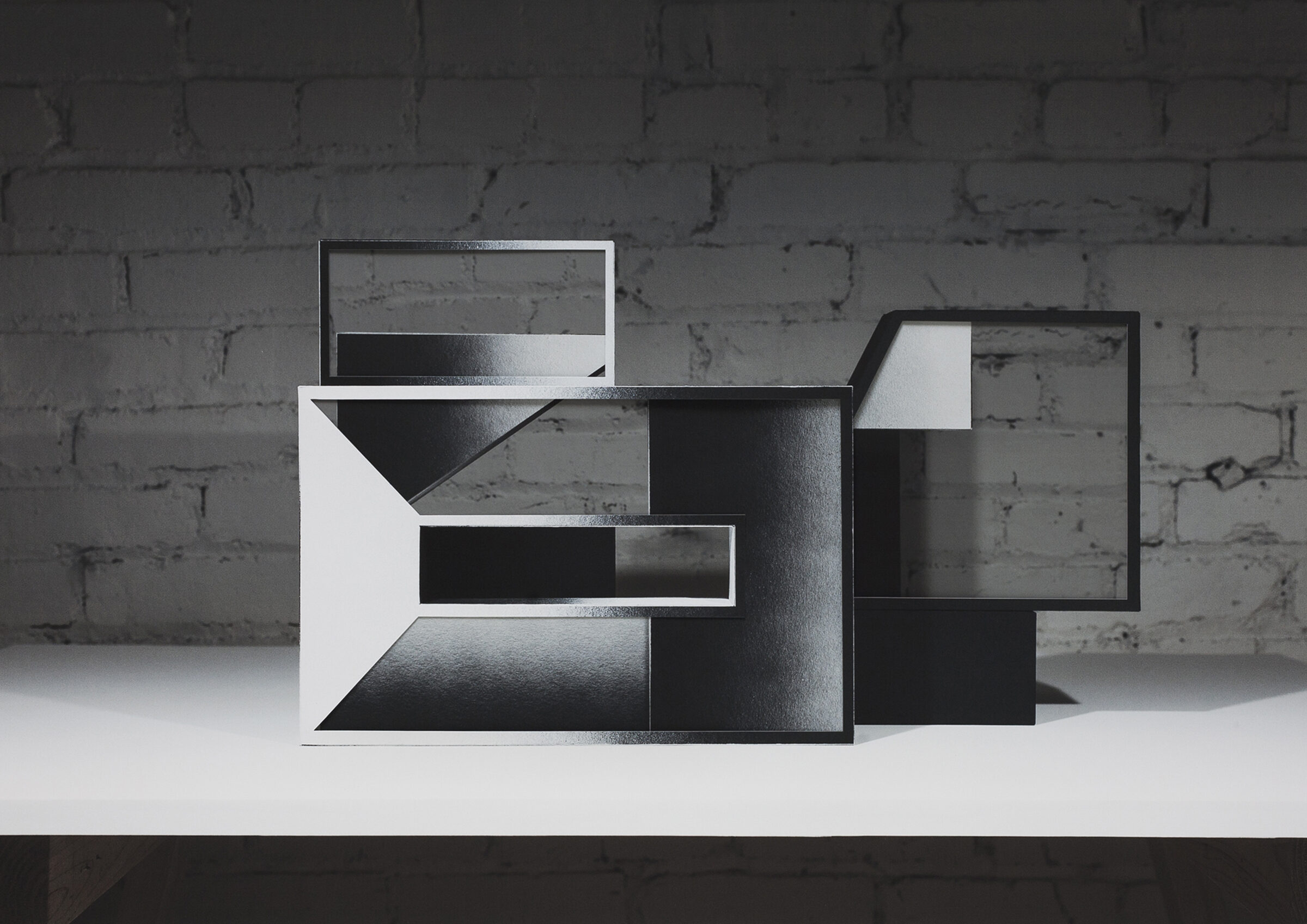 Abstract geometric composition featuring interlocking rectangular frames in black and white, layered to create a three-dimensional effect against a weathered brick wall background. The fragmented, overlapping planes suggest architectural forms with varying depths and contrasting tones.