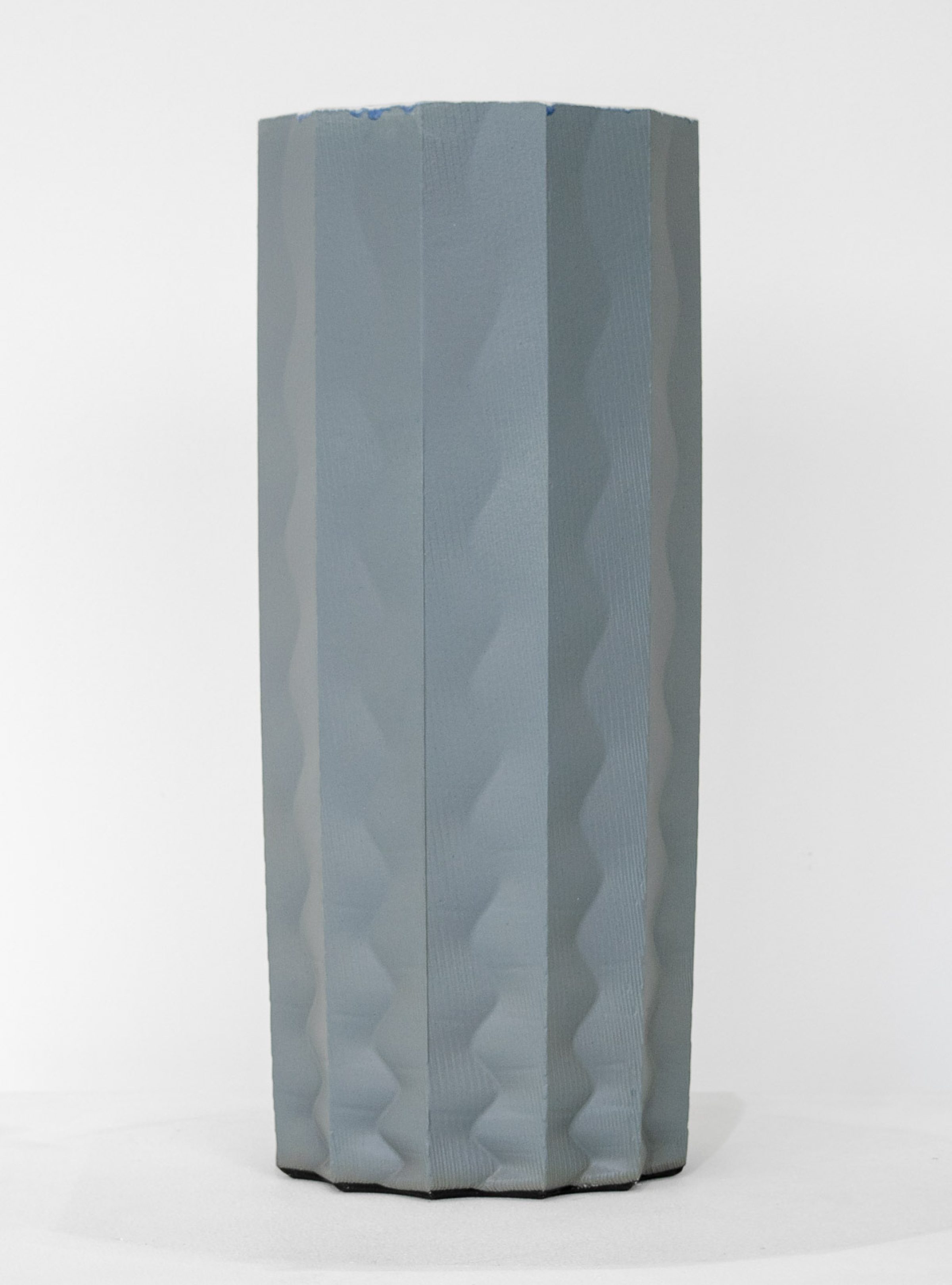 A tall cylindrical porcelain form with vertical ridged panels in soft gray and white tones, featuring subtle wave-like undulations across its surface. The sculpture stands against a neutral background, its organic, fluted structure creating areas of shadow and depth.