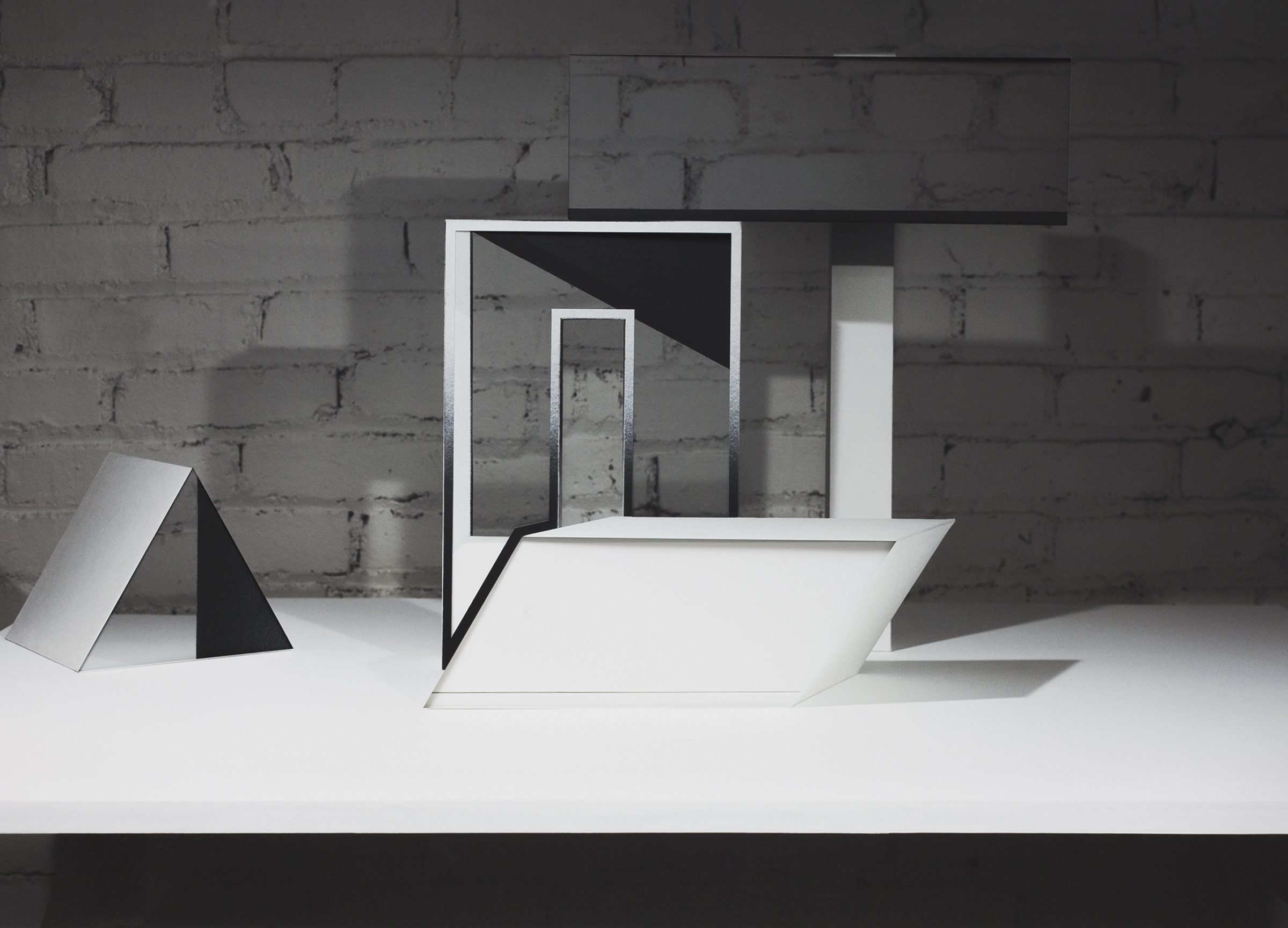 White and black geometric paper forms arranged on a white platform against a weathered brick wall. The sculptural composition features angular planes, rectangular frames, and triangular shapes in varying scales, creating an abstract architectural study in monochromatic tones.