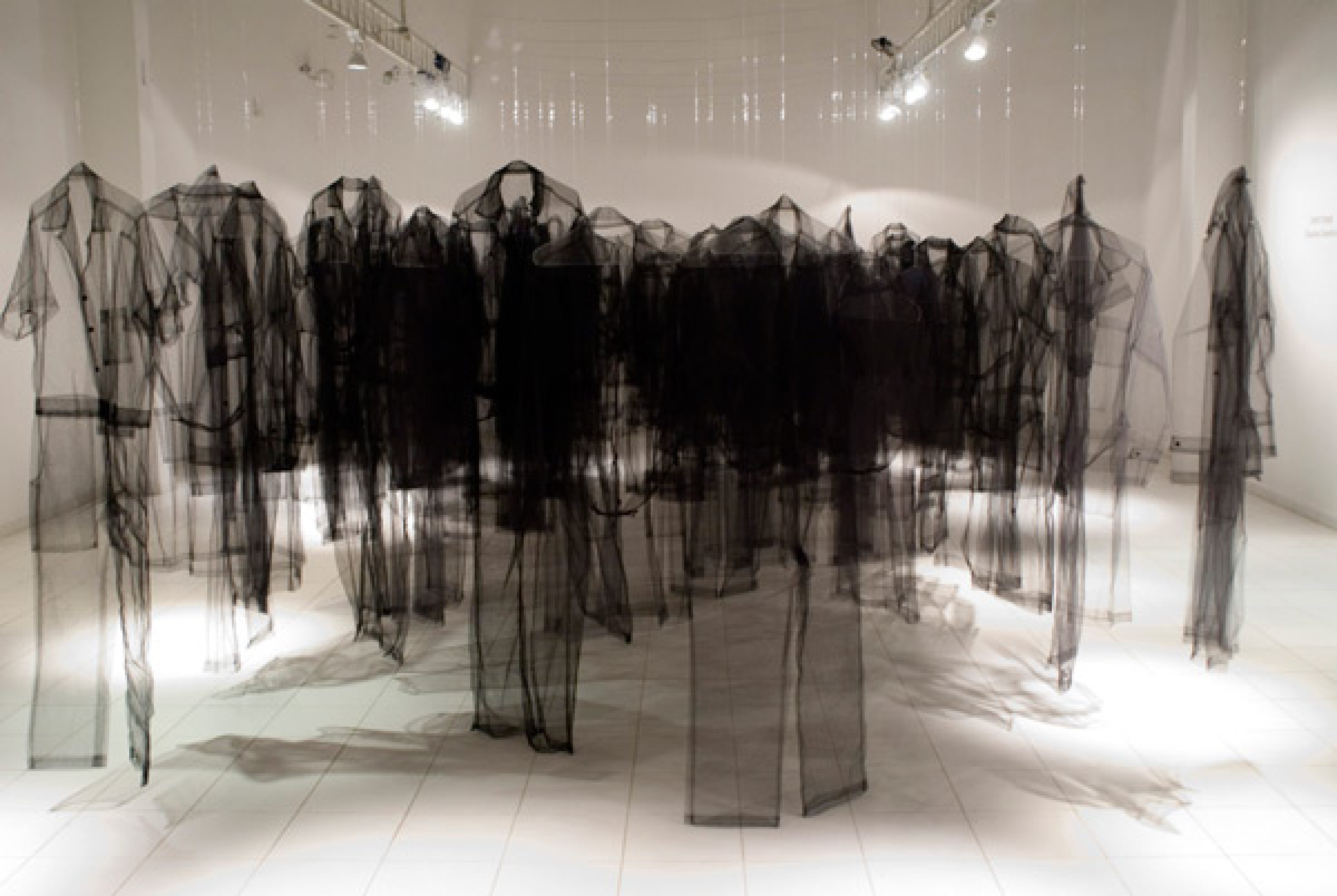 A large-scale room installation featuring numerous translucent black tulle garments suspended from above, creating silhouettes of human figures that overlap and layer throughout the white gallery space, with dramatic shadows cast by overhead lighting.