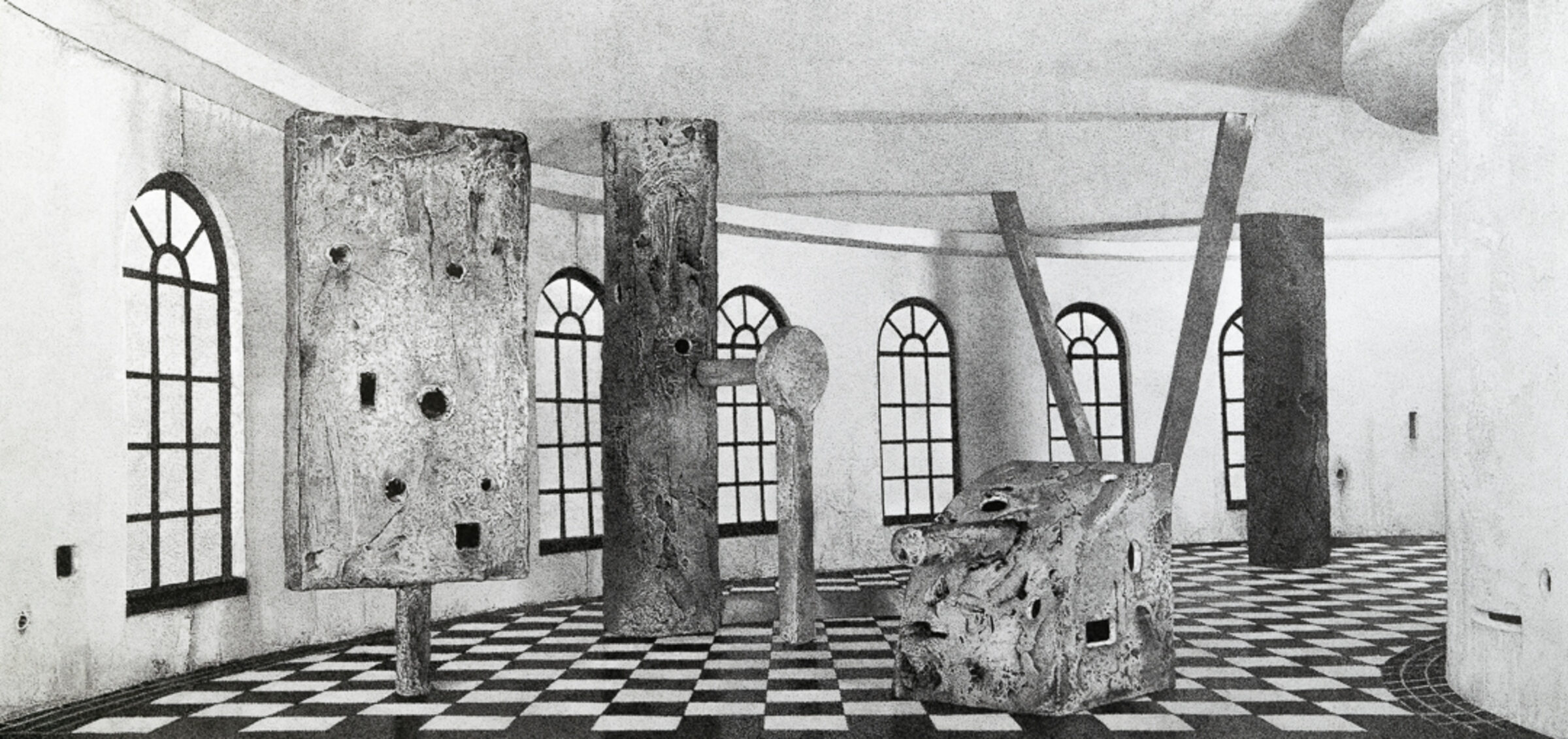 Black and white architectural interior with weathered concrete columns and walls featuring a geometric checkerboard floor. Multiple arched windows with grid patterns line the space, while irregular holes and openings punctuate the surfaces, creating an ambiguous sense of decay or deliberate abstraction.