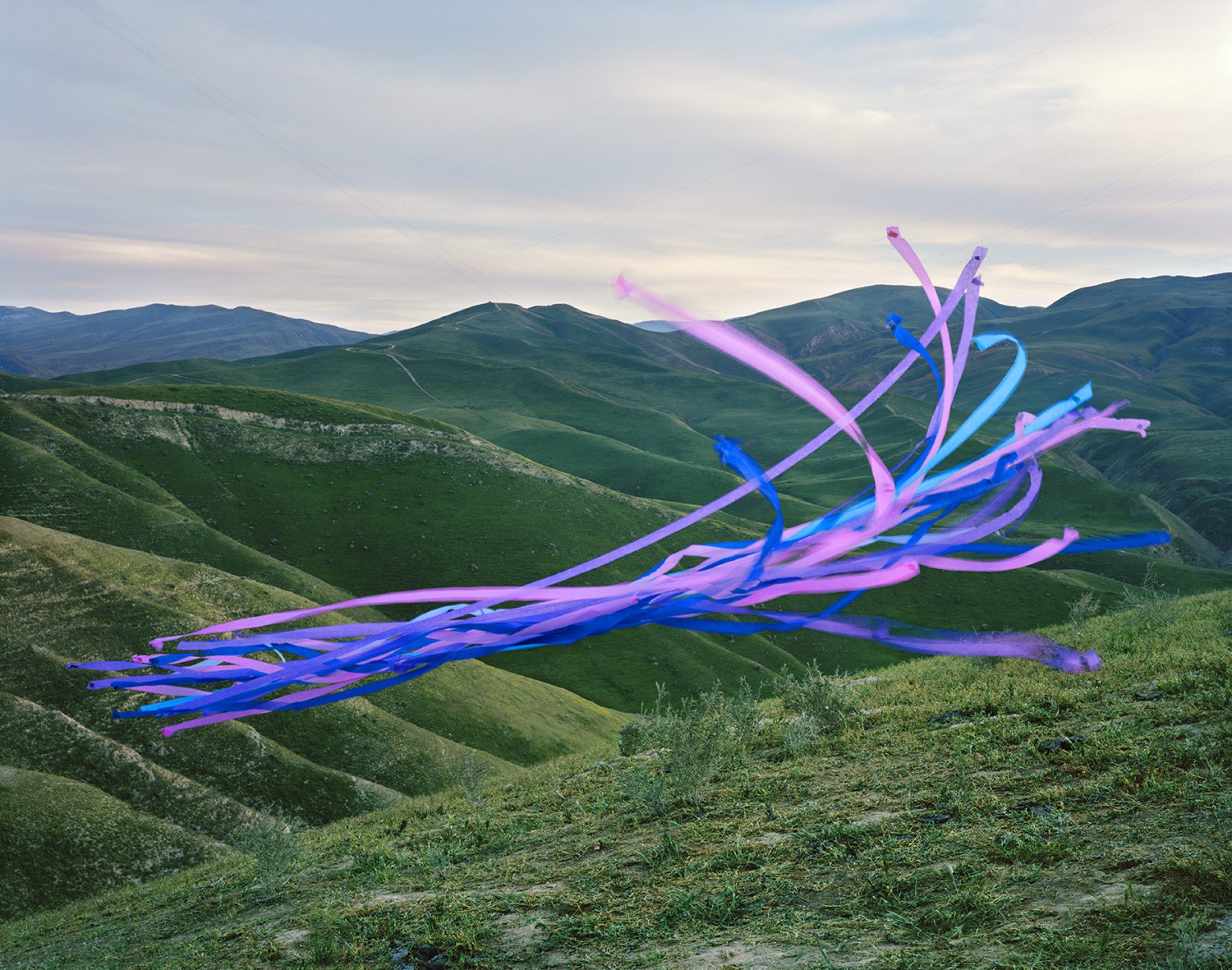 Vibrant neon-colored streamers in shades of blue, pink, and purple cascade across a rolling landscape of green hills under an overcast sky, creating a striking contrast between the digital-looking synthetic forms and the natural terrain.