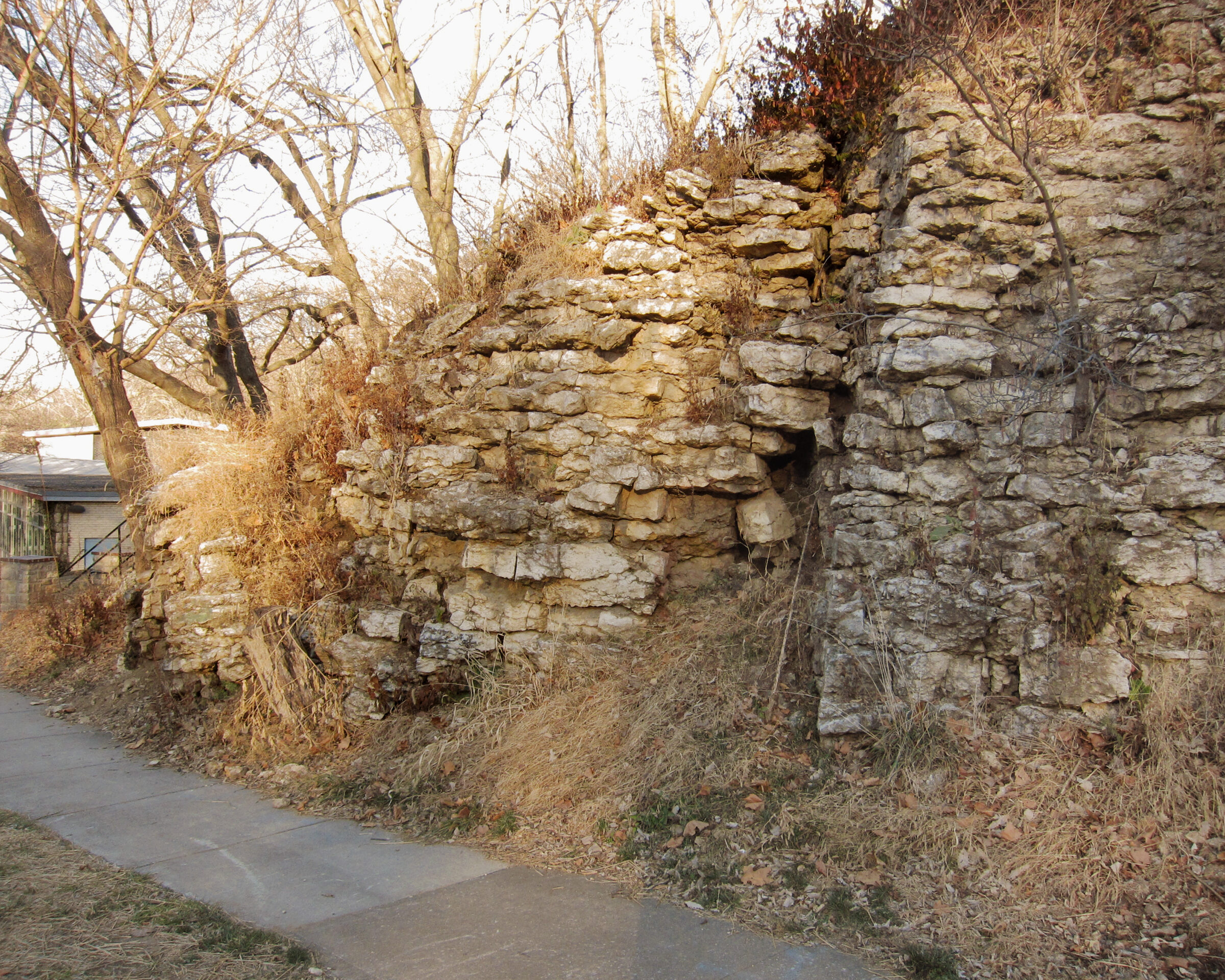 Stone ruins of stacked tan and grey limestone blocks form a tall wall alongside a concrete pathway, with bare winter trees and dried vegetation visible in the foreground and background.