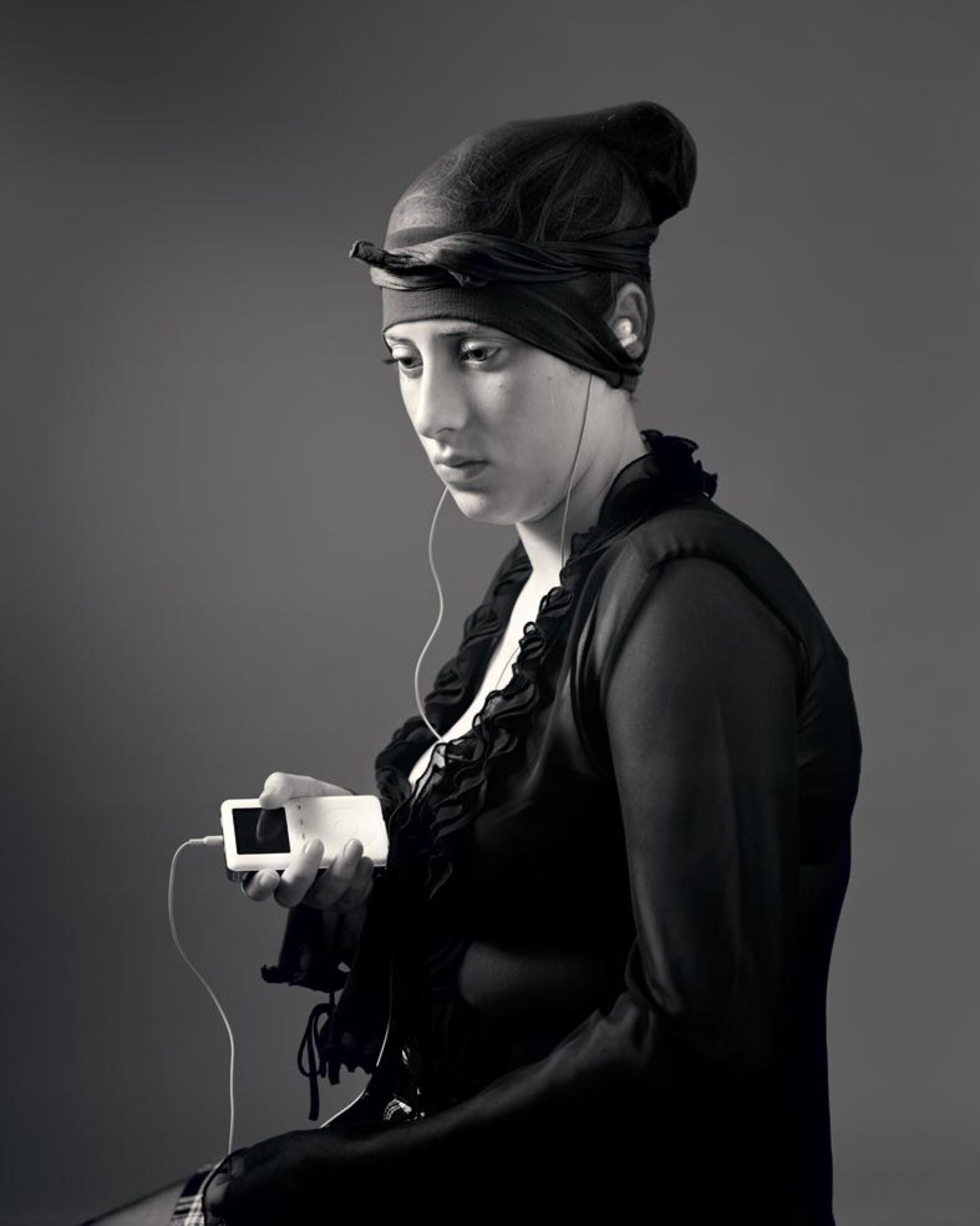 Black and white portrait of a woman in profile wearing a black hooded jacket and headband, holding a small white digital device with white earbuds. The minimalist composition is set against a neutral gray background with dramatic lighting that emphasizes the subject's contemplative expression.