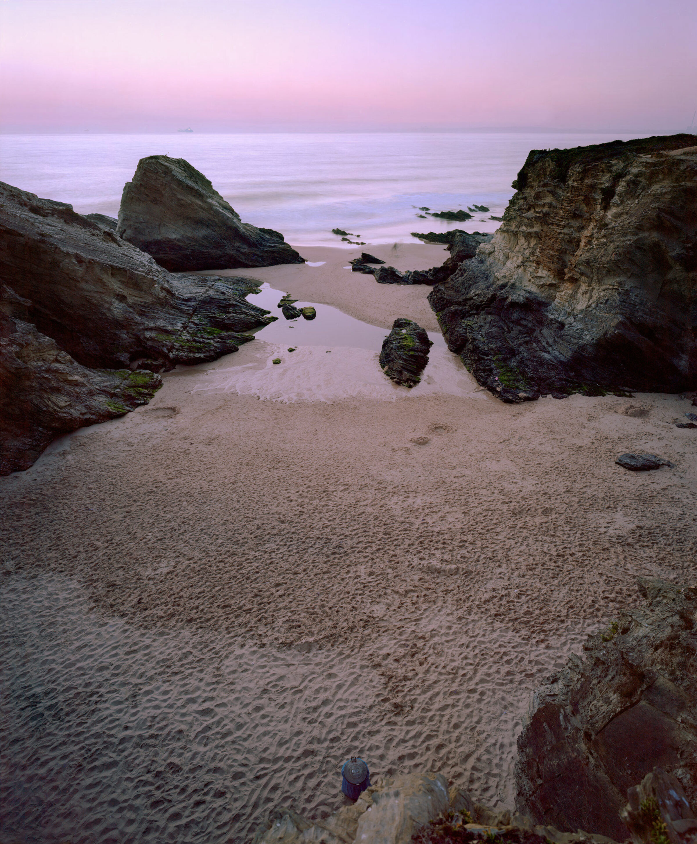 A coastal scene at dusk with dramatic rocky outcroppings framing a sandy beach and shallow pools of water. The sky glows in soft pink and lavender tones, while moss-covered stones and weathered rock formations create a sense of geological depth and layering across the composition.