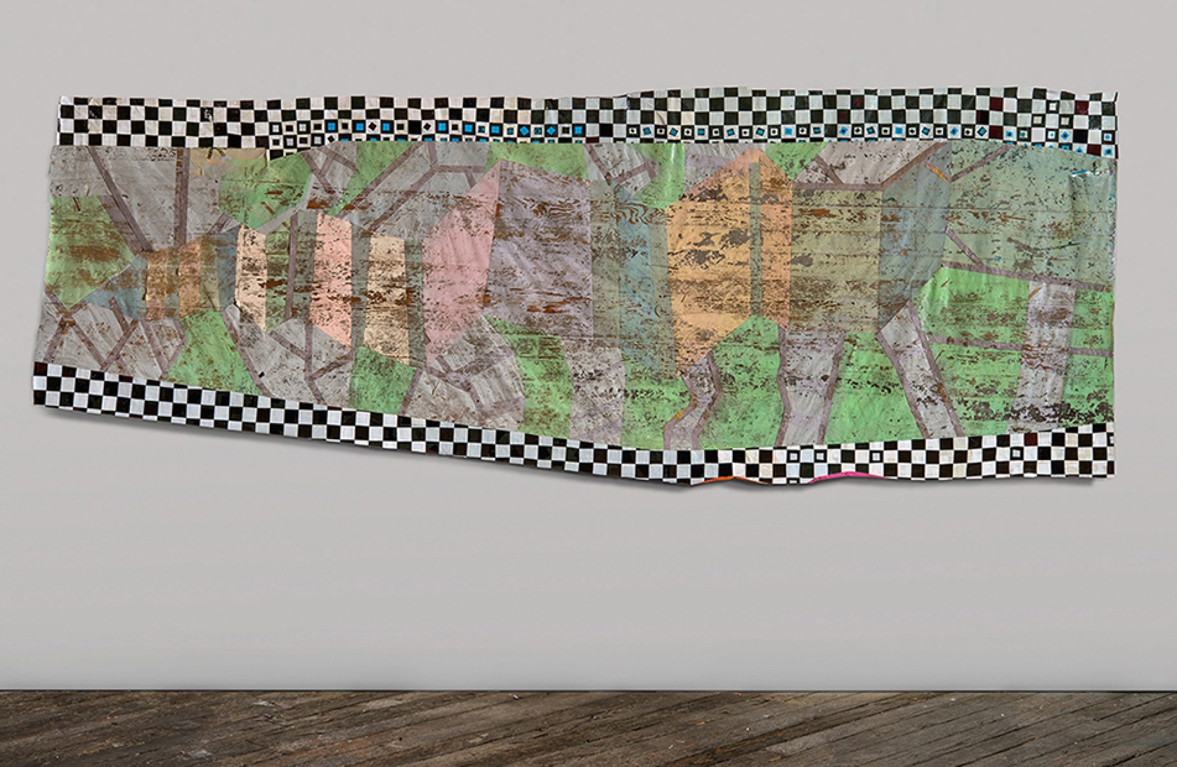 A horizontal mixed-media collage featuring layered aerial or cartographic imagery in greens, browns, and muted tones, bordered by a black and white checkered pattern. The composition is mounted on a light gray wall above wooden flooring, with the irregular edges and translucent quality of the materials creating depth and visual complexity.