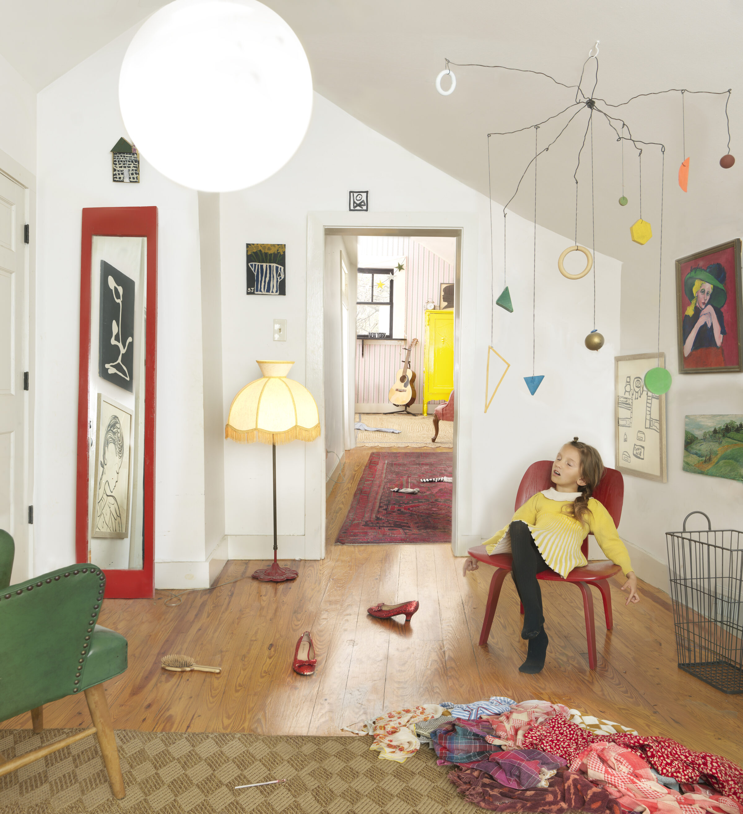 A young girl in a yellow sweater sits in a burgundy chair within a bright, eclectic interior space. The room features white walls, wooden flooring, and is filled with colorful artworks, hanging mobiles with painted geometric shapes and spheres, a yellow-shaded floor lamp, and scattered red shoes on the floor, creating a whimsical, creative domestic environment.