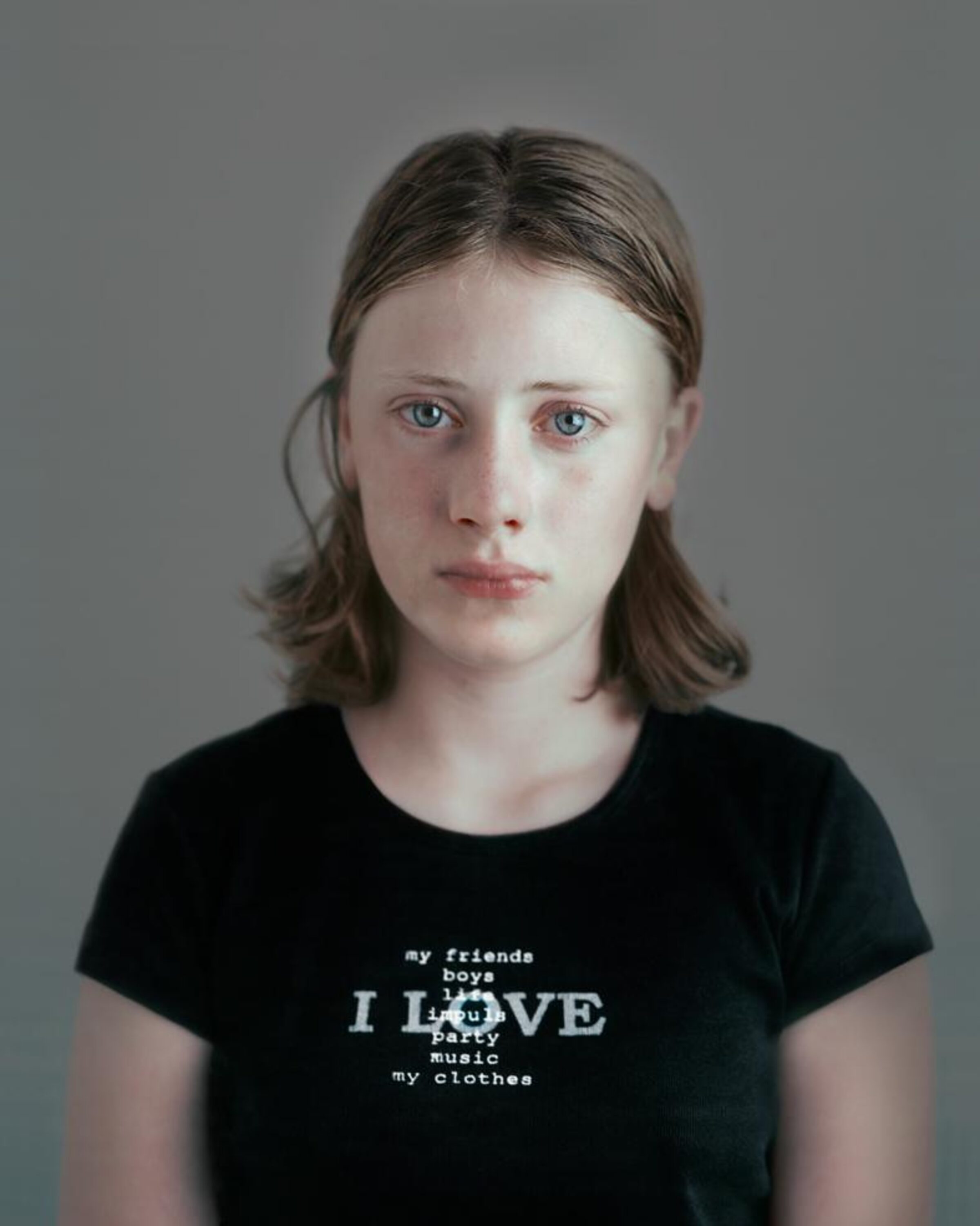 Portrait of a young person with shoulder-length brown hair wearing a black t-shirt printed with white text listing "my friends boys I LOVE party music my clothes" against a neutral gray background. The photograph employs a direct, frontal composition with a contemplative expression and cool color palette.