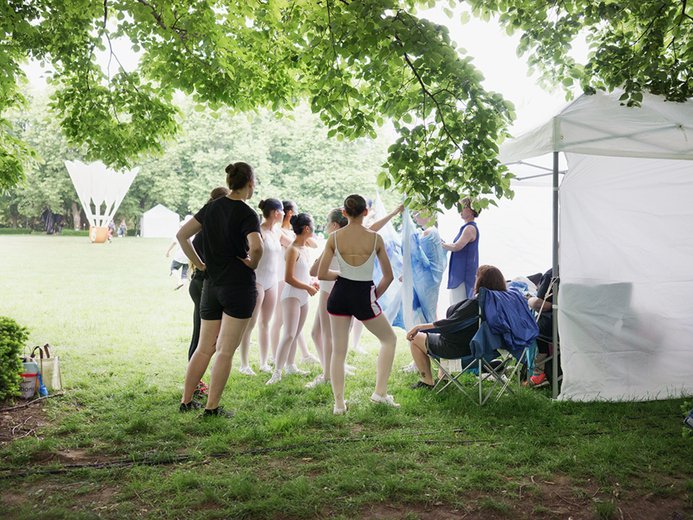 A group of young dancers in ballet attire gather beneath leafy trees in a grassy park setting, with a white tent and spectators visible in the background. The scene captures a moment of community and performance in an outdoor natural environment.