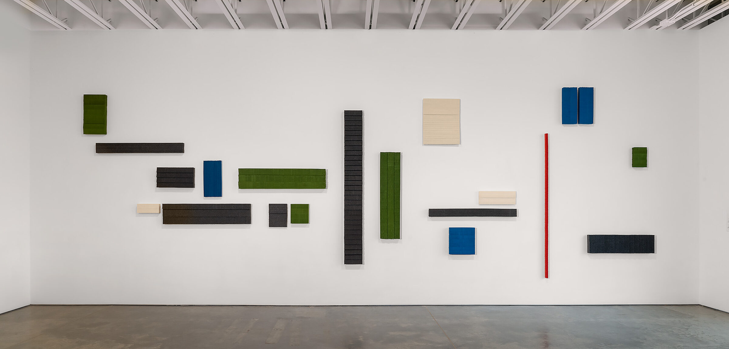 Abstract wall installation composed of rectangular and square panels in varying sizes arranged across a white gallery wall. Panels in dark charcoal, forest green, cream, blue, and a single red vertical element create a rhythmic composition against the minimalist white backdrop.