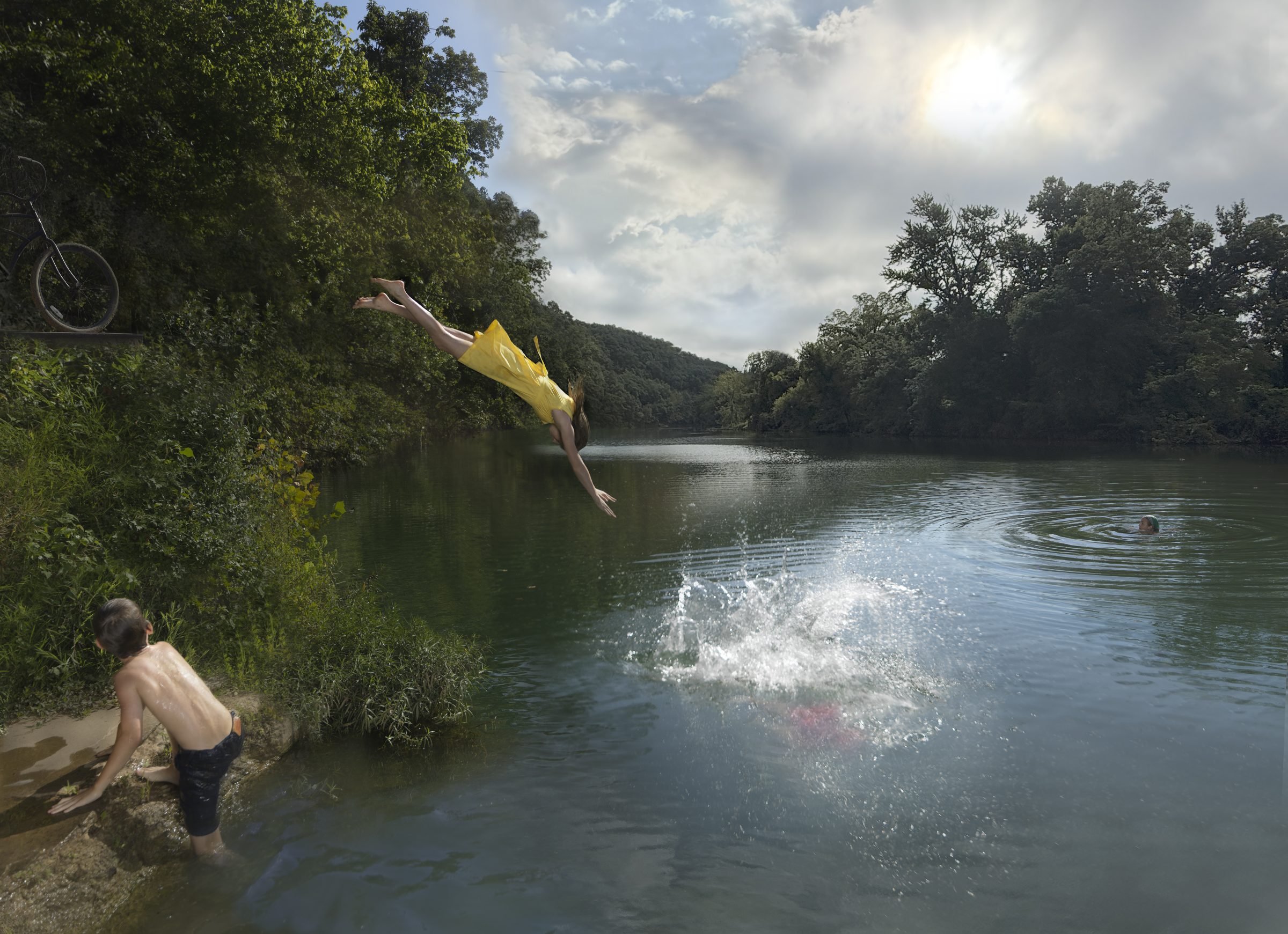 A person in a bright yellow shirt mid-dive from a riverbank toward dark green water, captured in dynamic motion with arms outstretched. Another swimmer watches from the shallow shore below overhanging vegetation, while a third figure swims in the distance, framed by forested hills and a cloudy sky.
